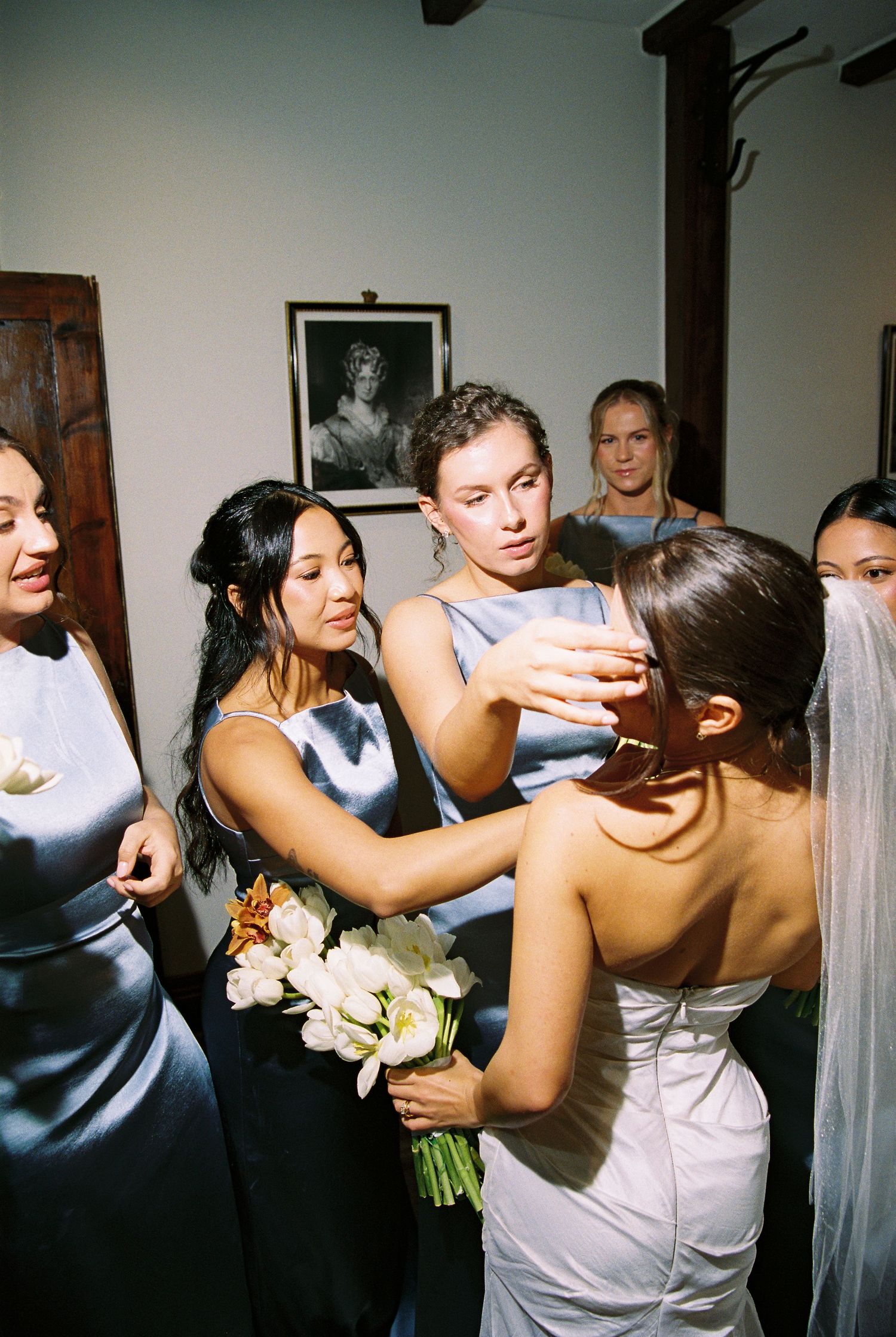 Bridesmaids in blue satin dresses gather around the bride adjusting her veil before ceremony.