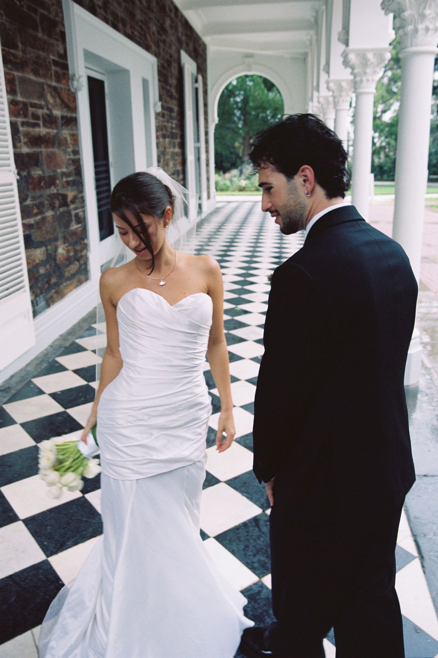 A bride in a strapless white mermaid gown and groom in a black suit walk along a checkered porch together.