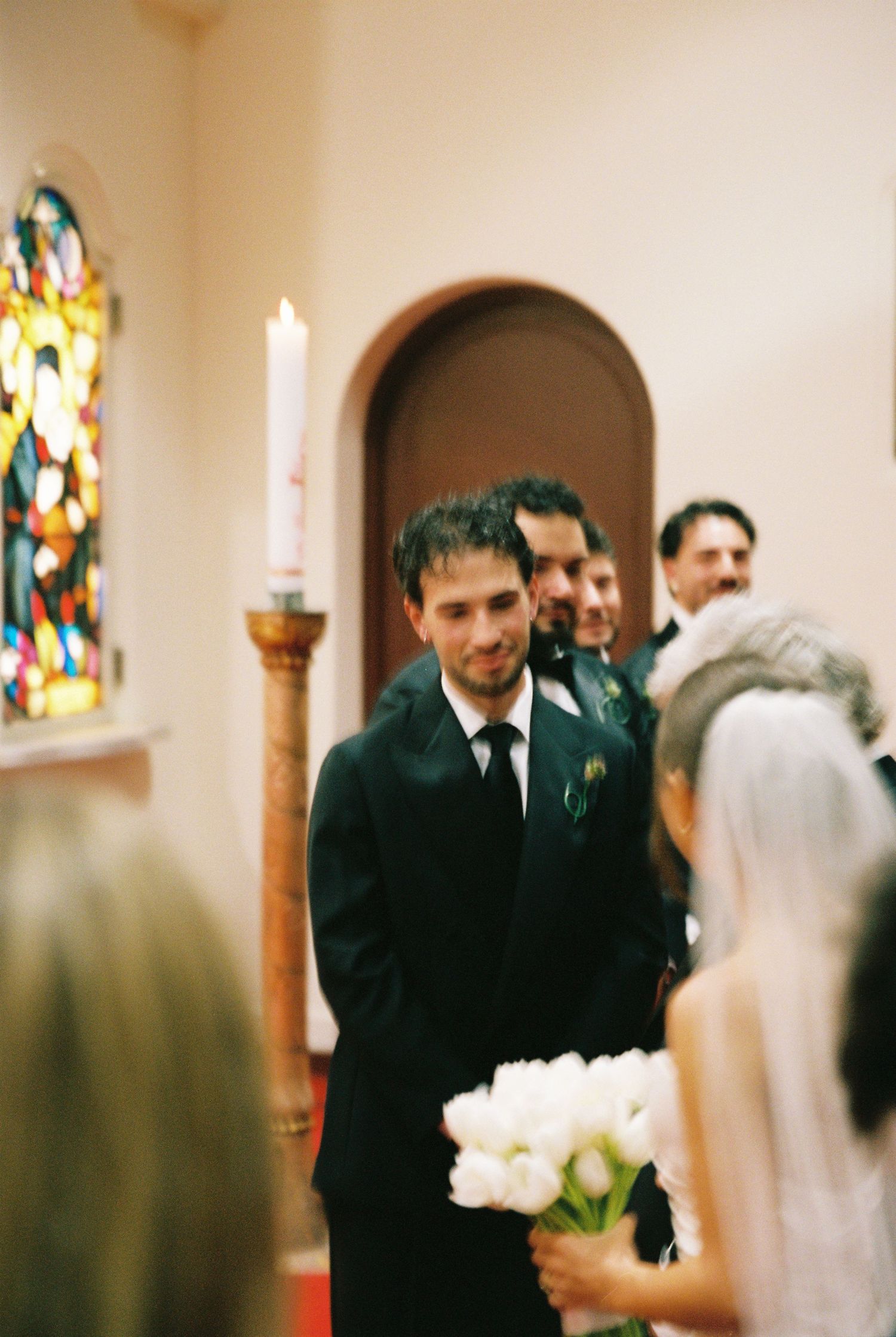 Groom in black suit smiling at bride walking down church aisle during wedding ceremony.