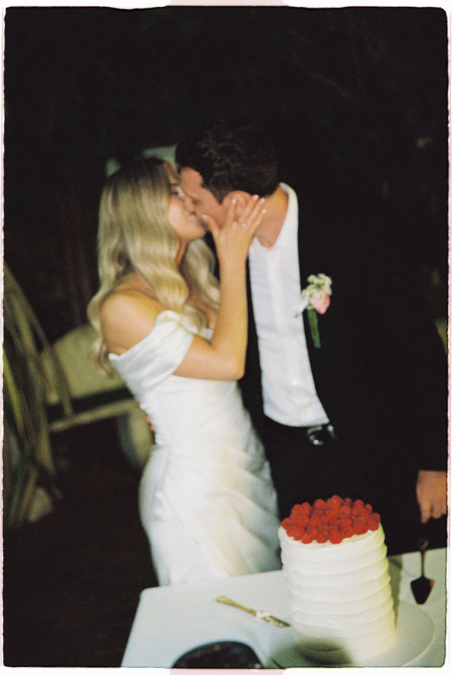 Newlyweds kiss behind white wedding cake topped with fresh red raspberries.