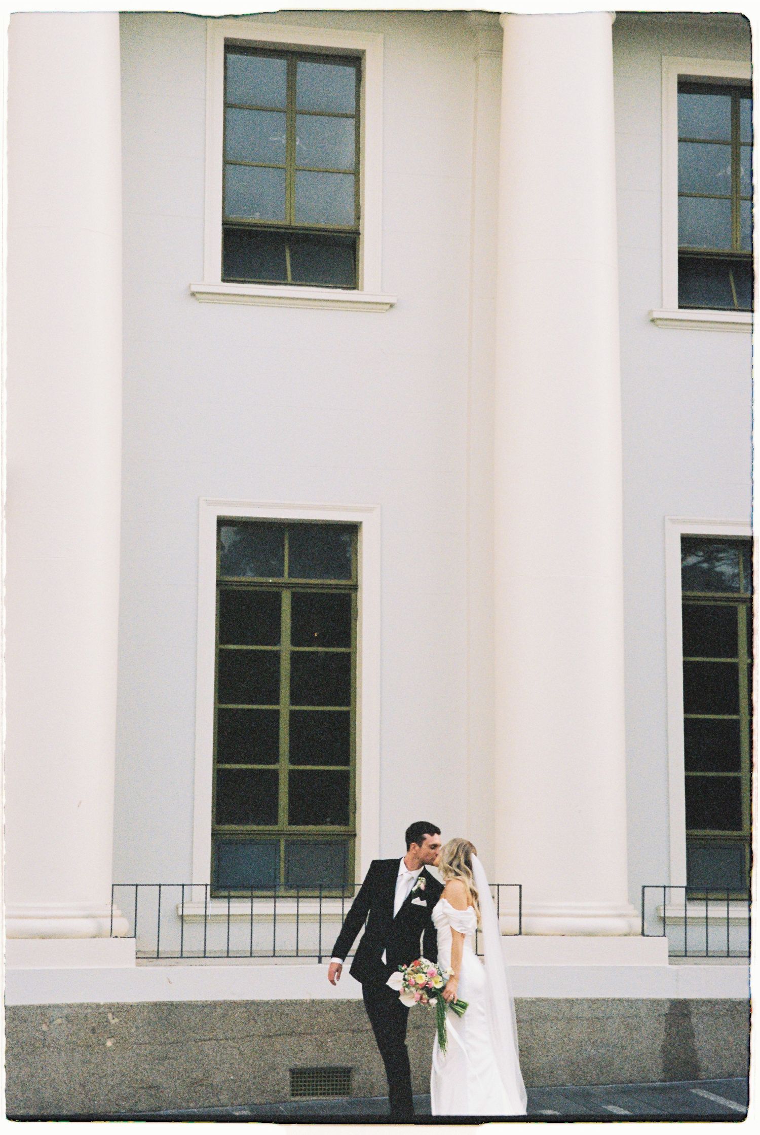 Bride and groom share a tender kiss beside tall white columns of an elegant building on their wedding day.