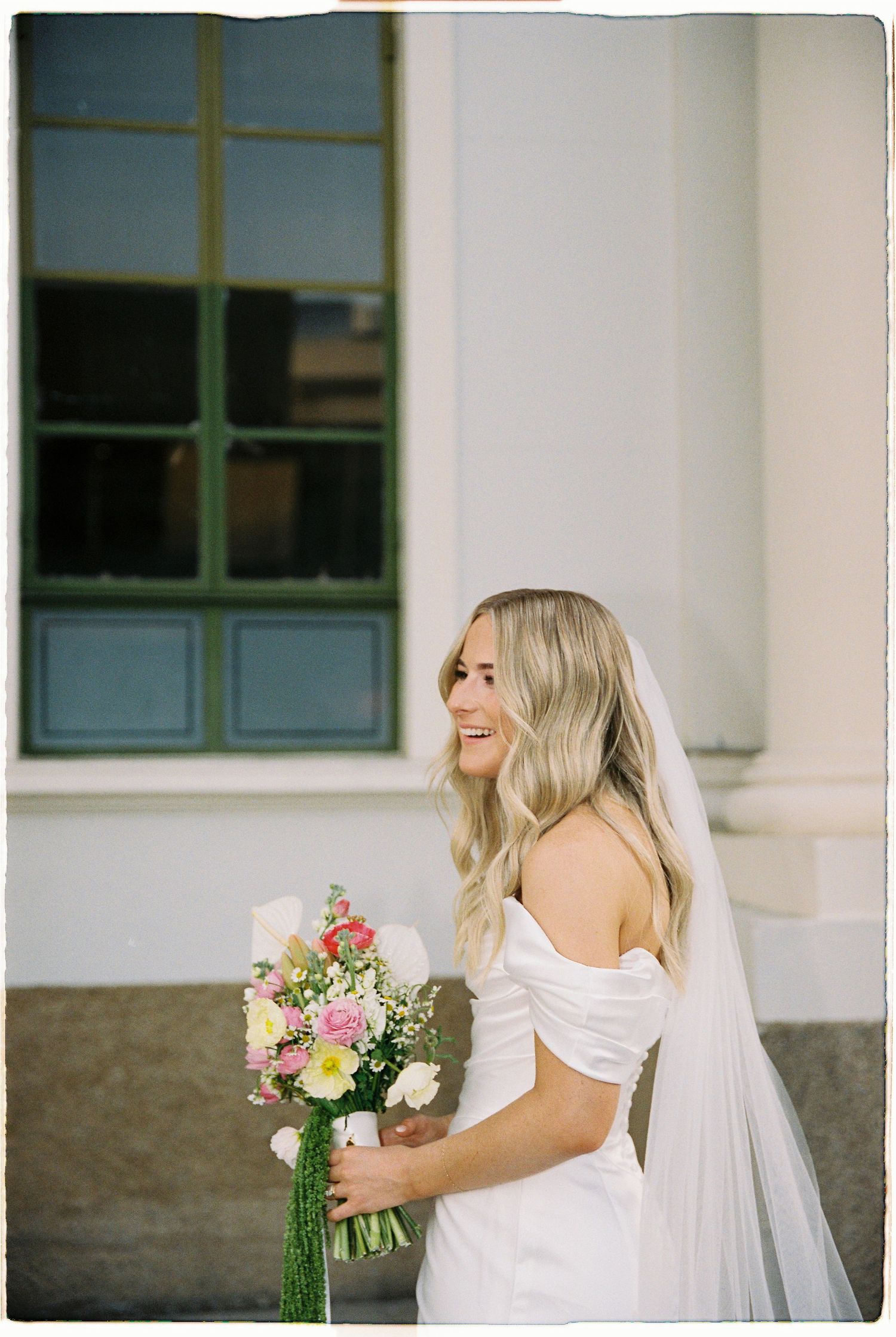 Laughing bride in off-shoulder white gown with veil holds colorful bouquet outside white classical building.