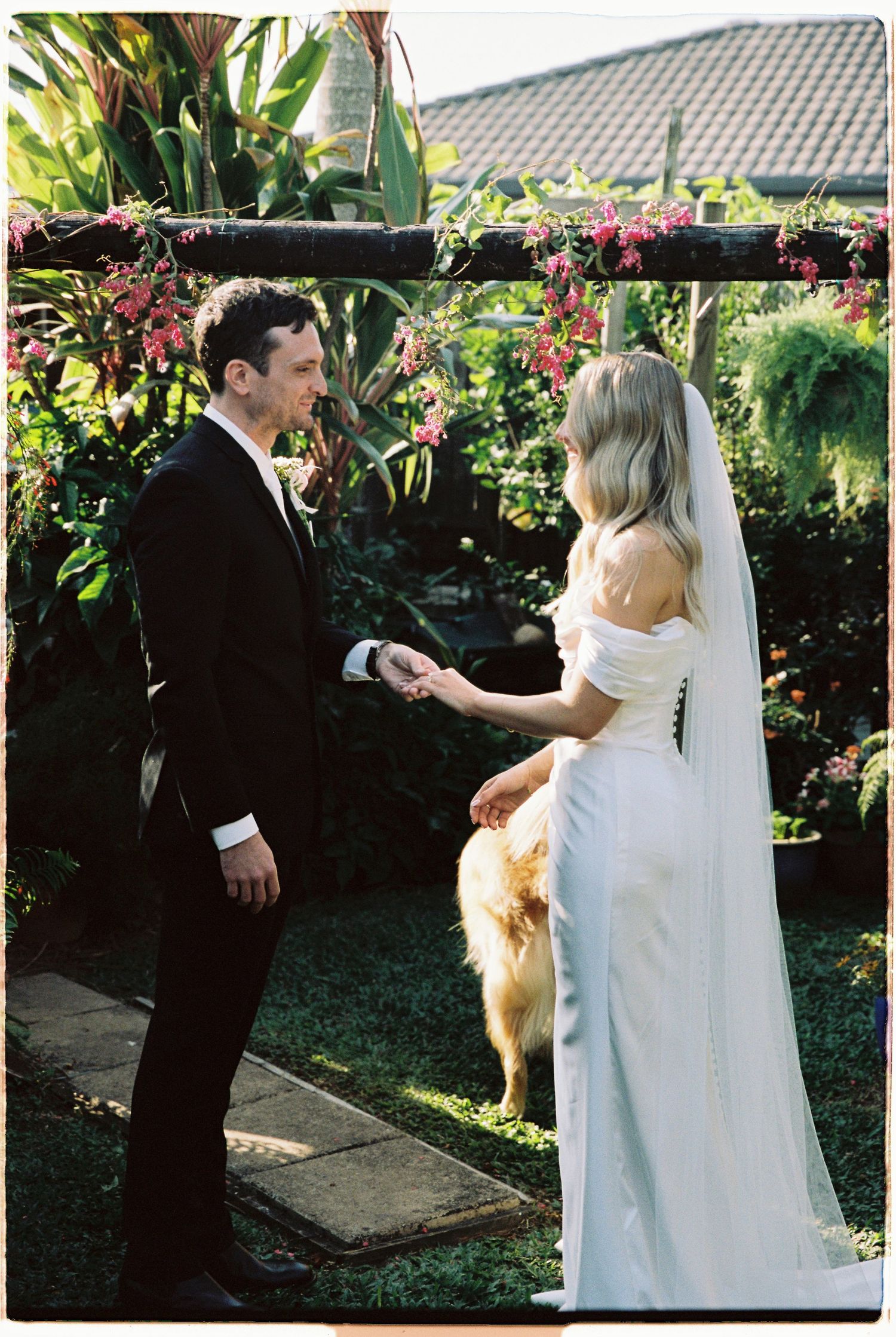 Bride and groom hold hands during outdoor garden wedding ceremony with lush tropical greenery and floral arbor.