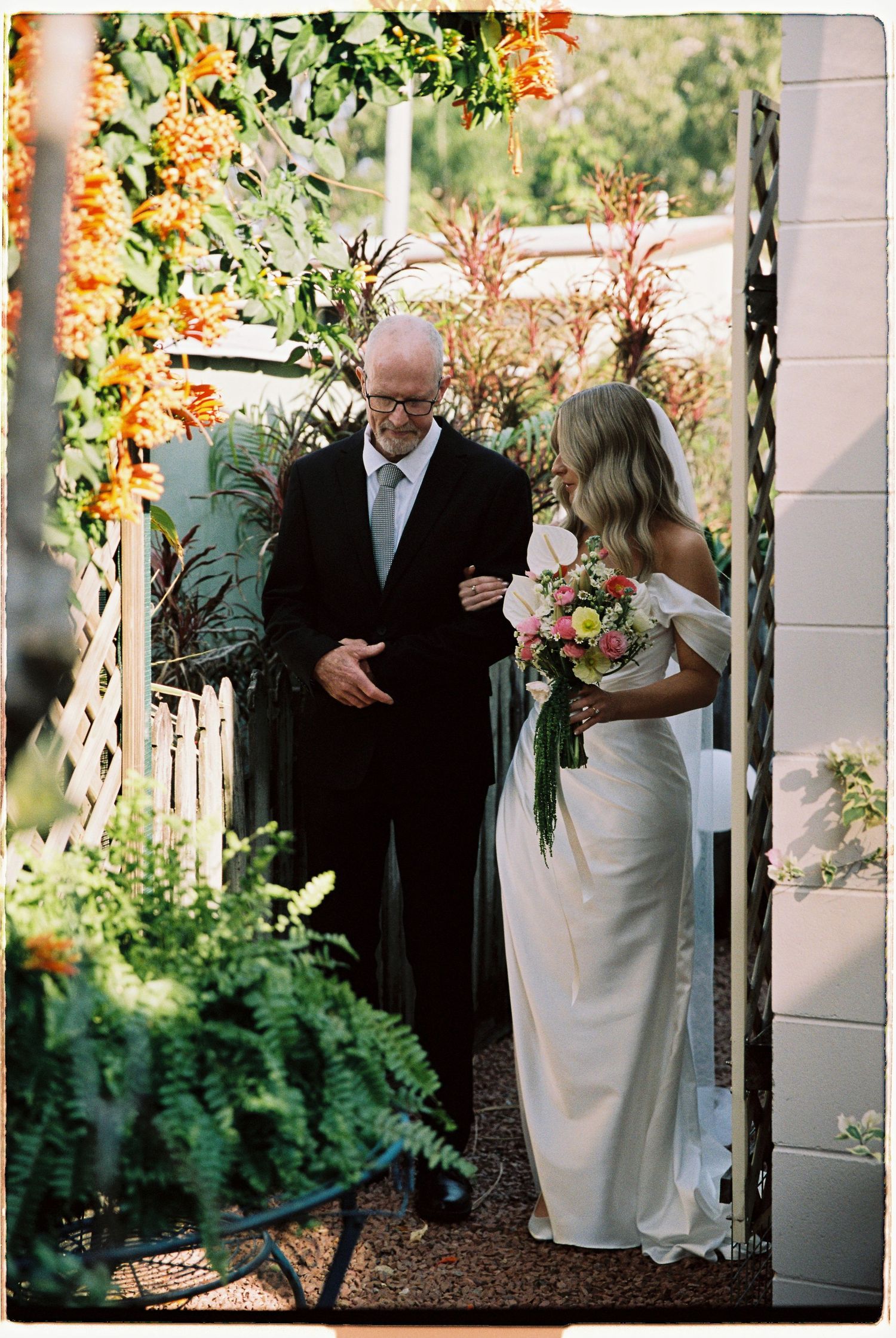 Bride walking with older man through floral archway at outdoor garden wedding.