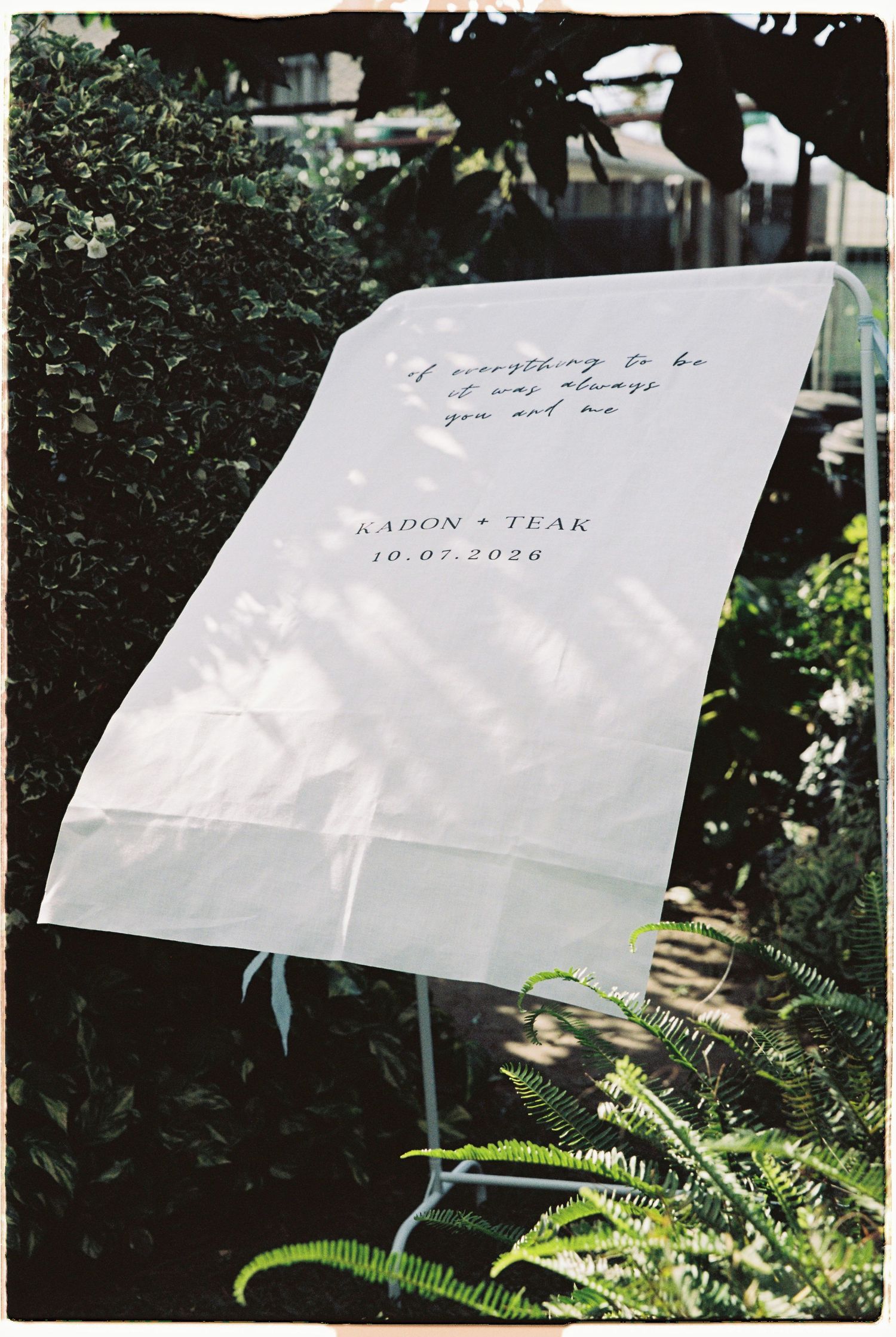 Elegant white wedding welcome sign reading KADON and TEAK 10.07.2026 displayed on stand in garden.