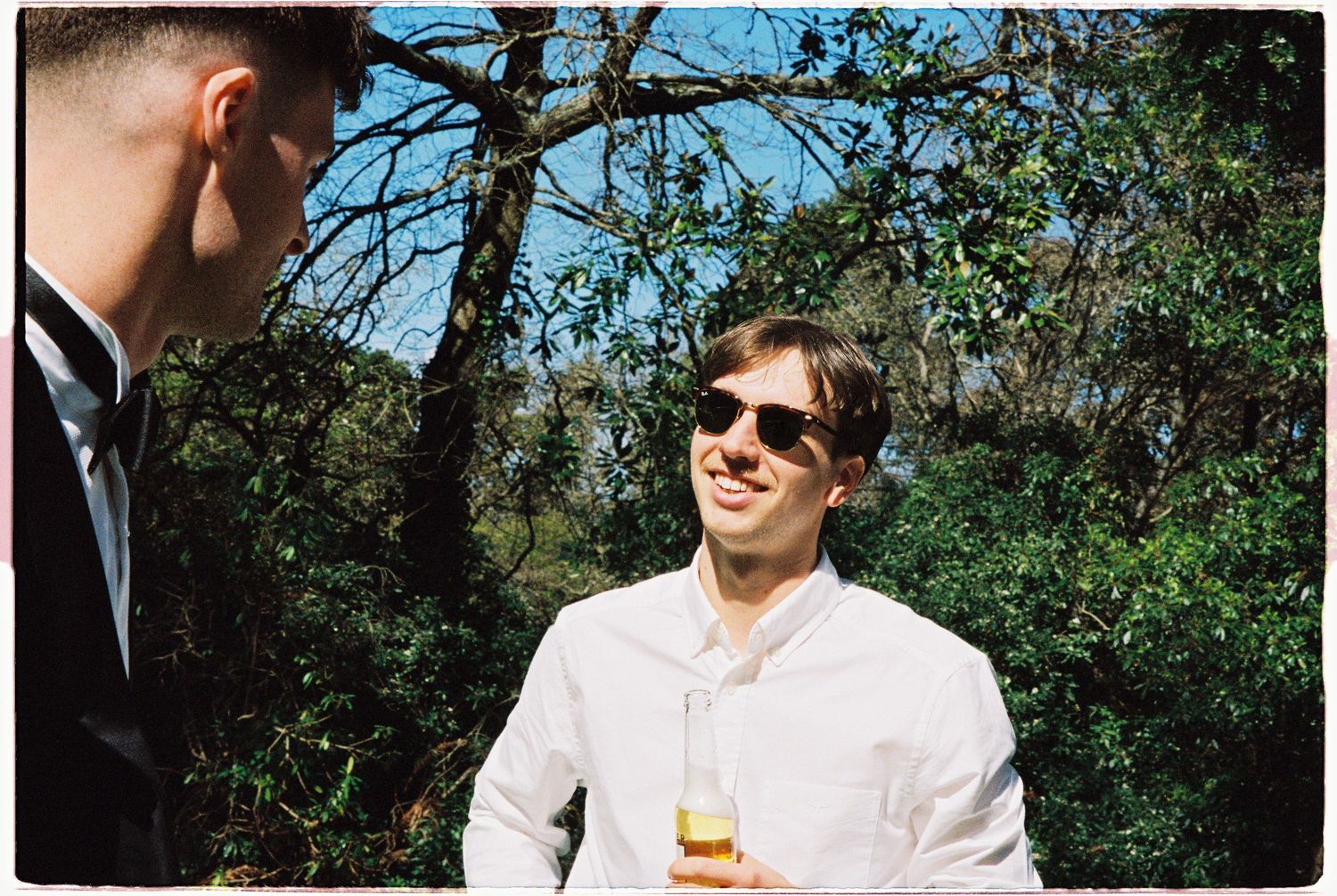 Two men in formal attire socializing outdoors, one holding a beer bottle, surrounded by lush green trees on a sunny day.