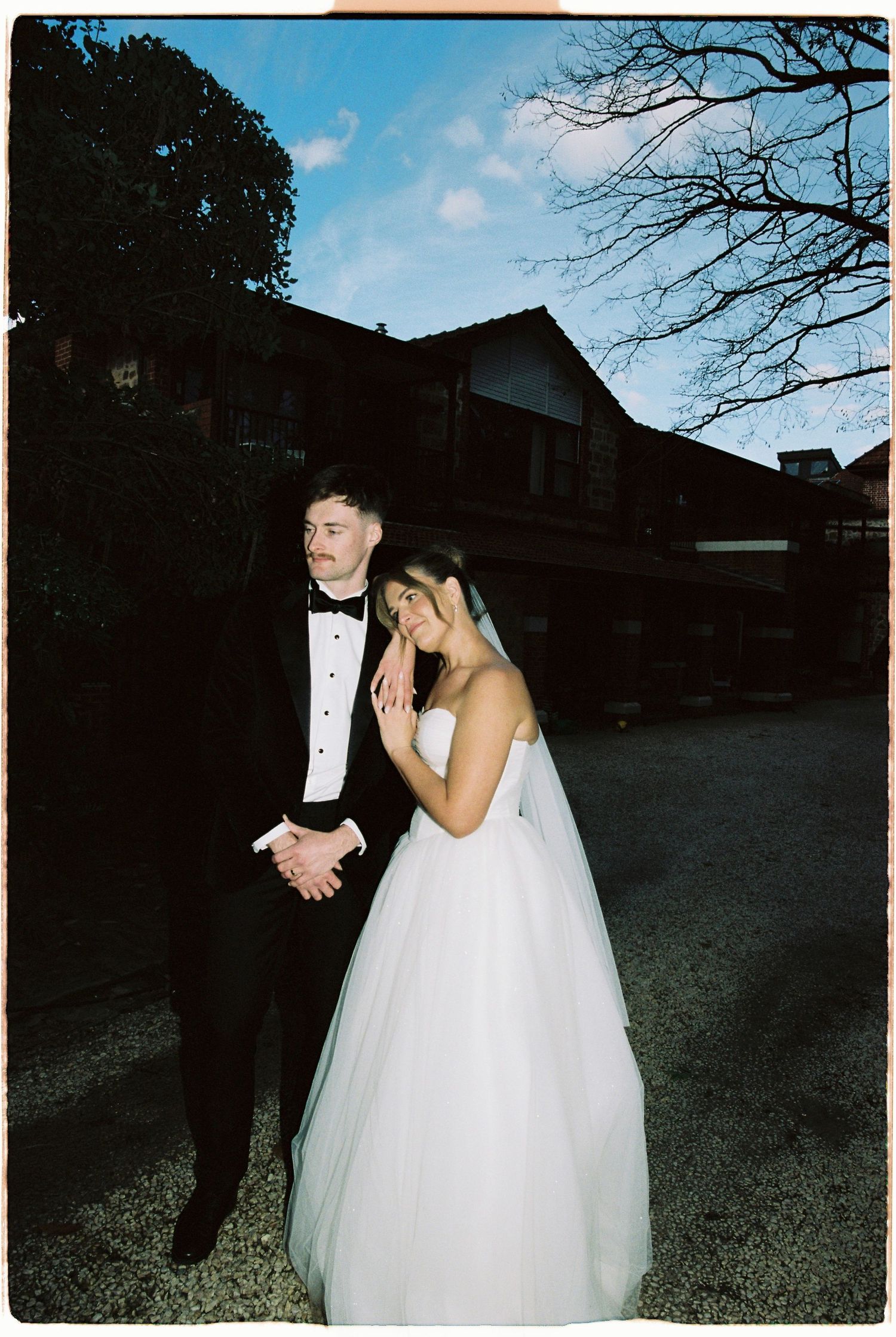 Bride in white ball gown and groom in black tuxedo posing together at dusk outside wedding venue.