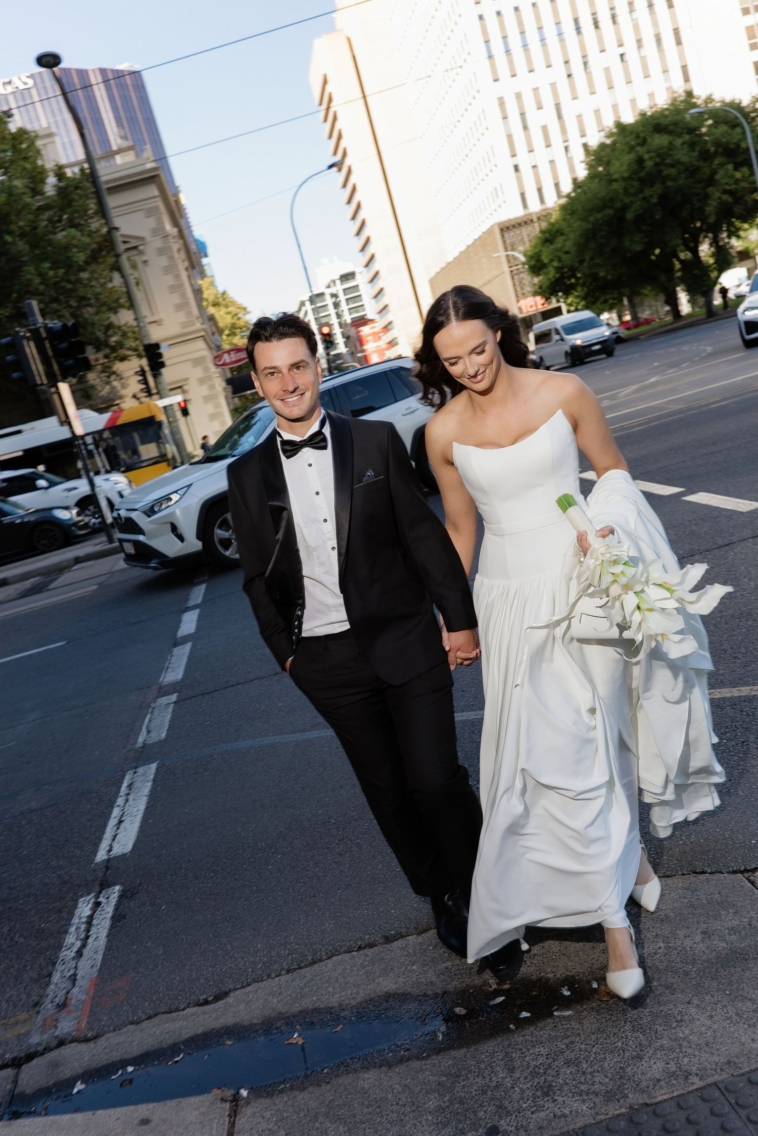 Smiling bride and groom holding hands walking across city street, bride in white strapless gown, groom in black tuxedo.
