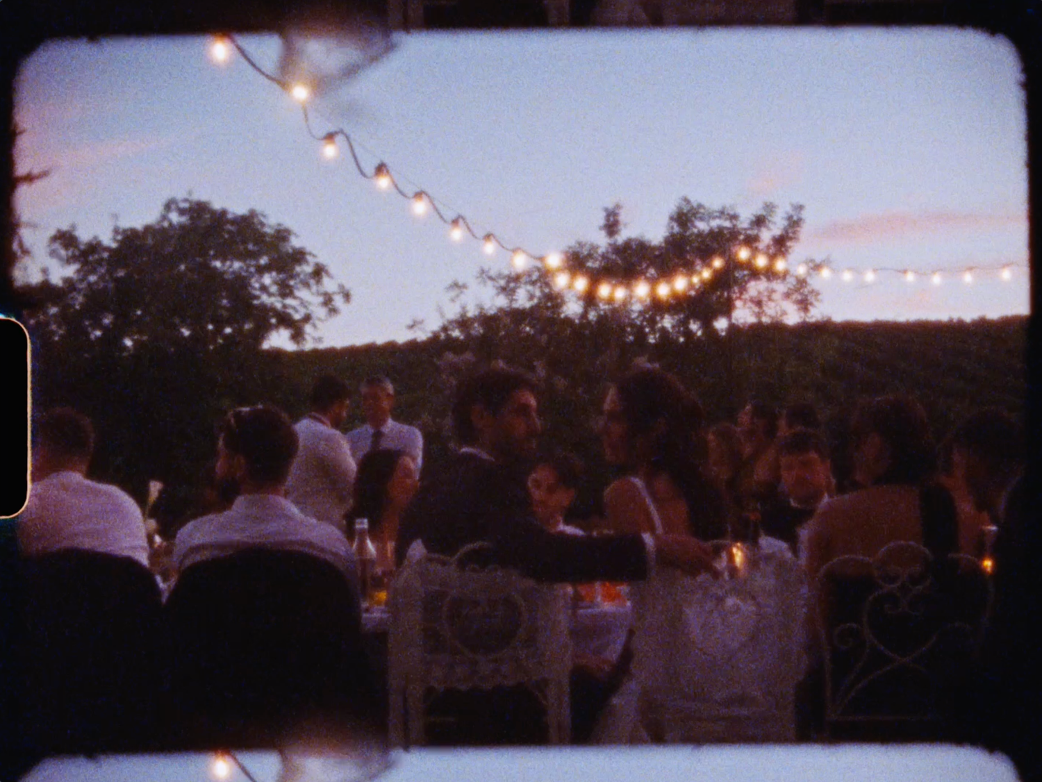 Guests dining at elegant outdoor wedding reception under string lights at dusk.