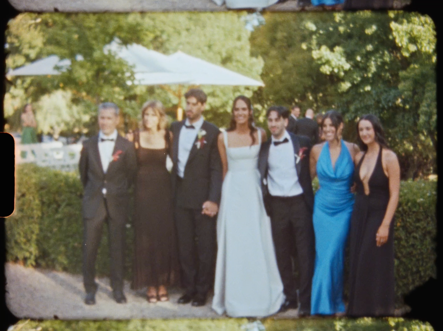 Wedding party posing in formal attire including a blue gown in a sunlit garden with green hedges.