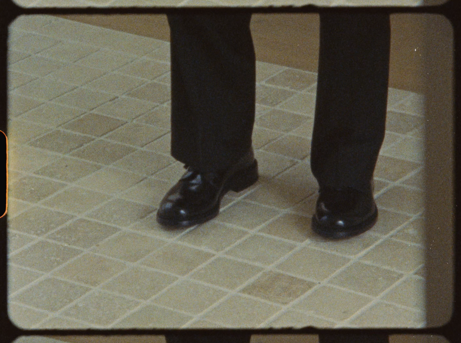 Close-up of a man's black dress shoes and dark trousers standing on a beige tile floor.