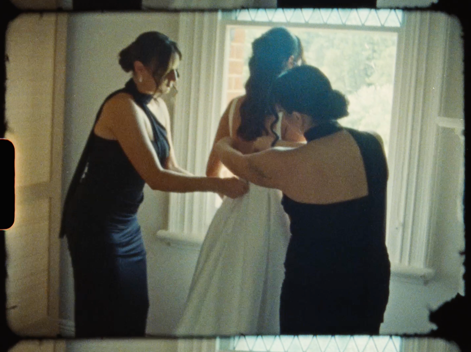 Two bridesmaids in navy dresses help a bride into her white wedding gown near a bright window.