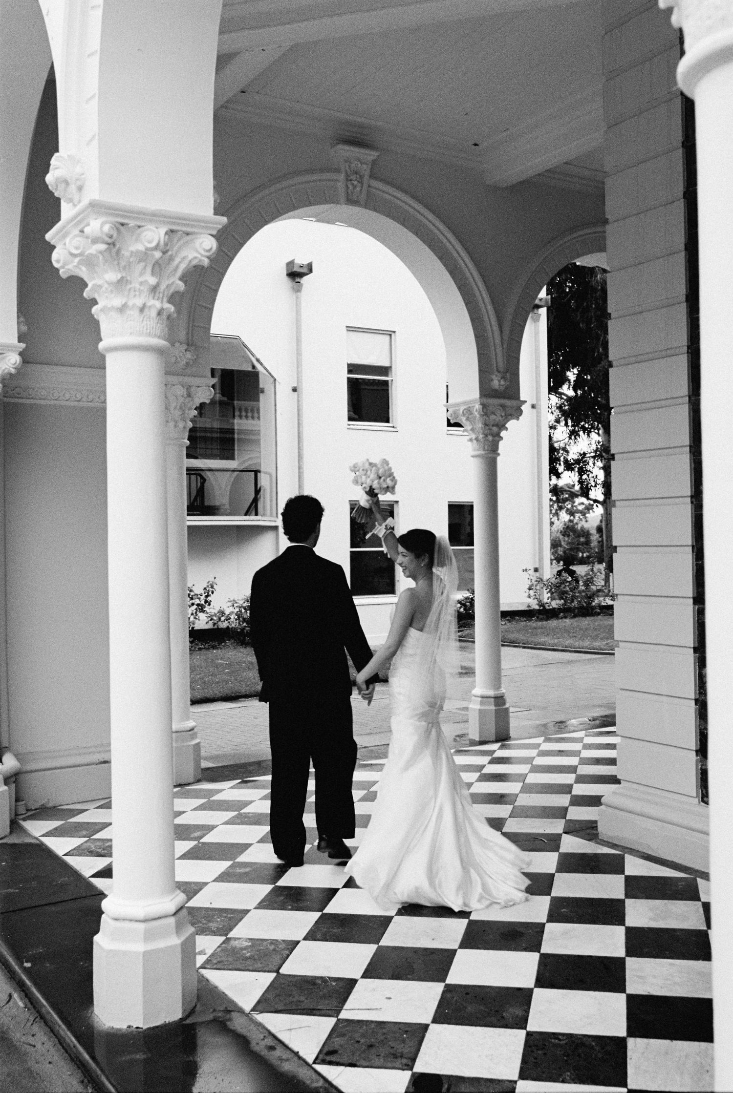Bride and groom hold hands under elegant arched colonnade with black and white checkered floor.
