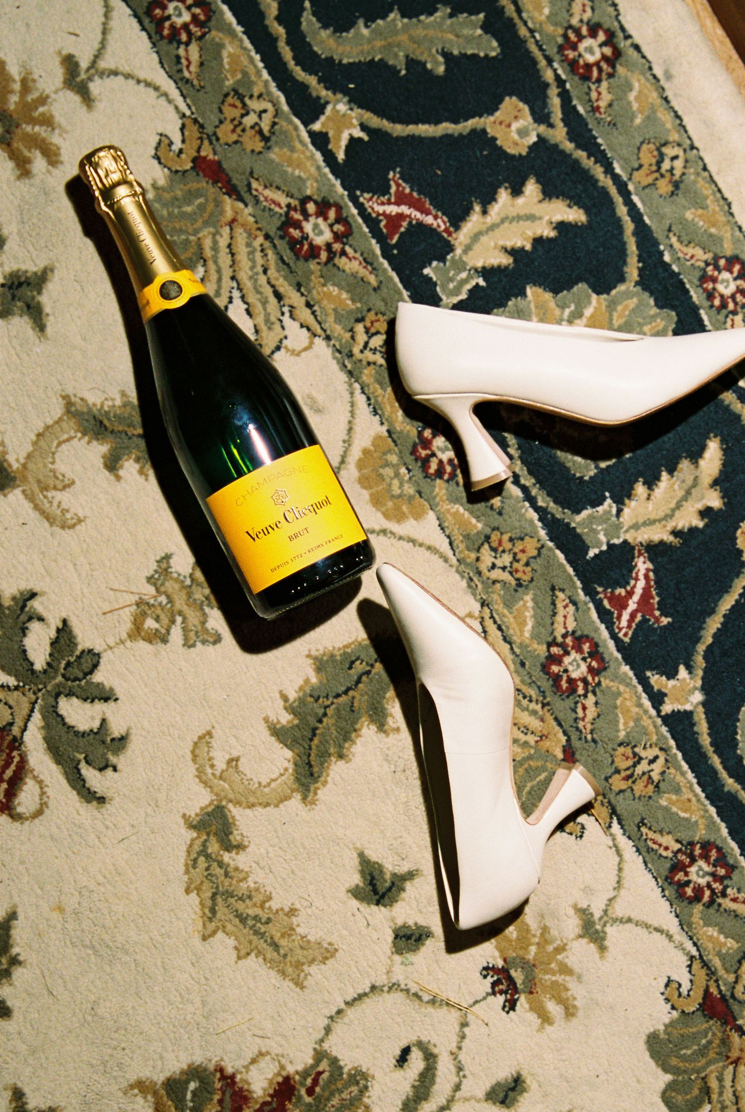 Veuve Clicquot champagne bottle beside white bridal heels on a floral patterned rug.