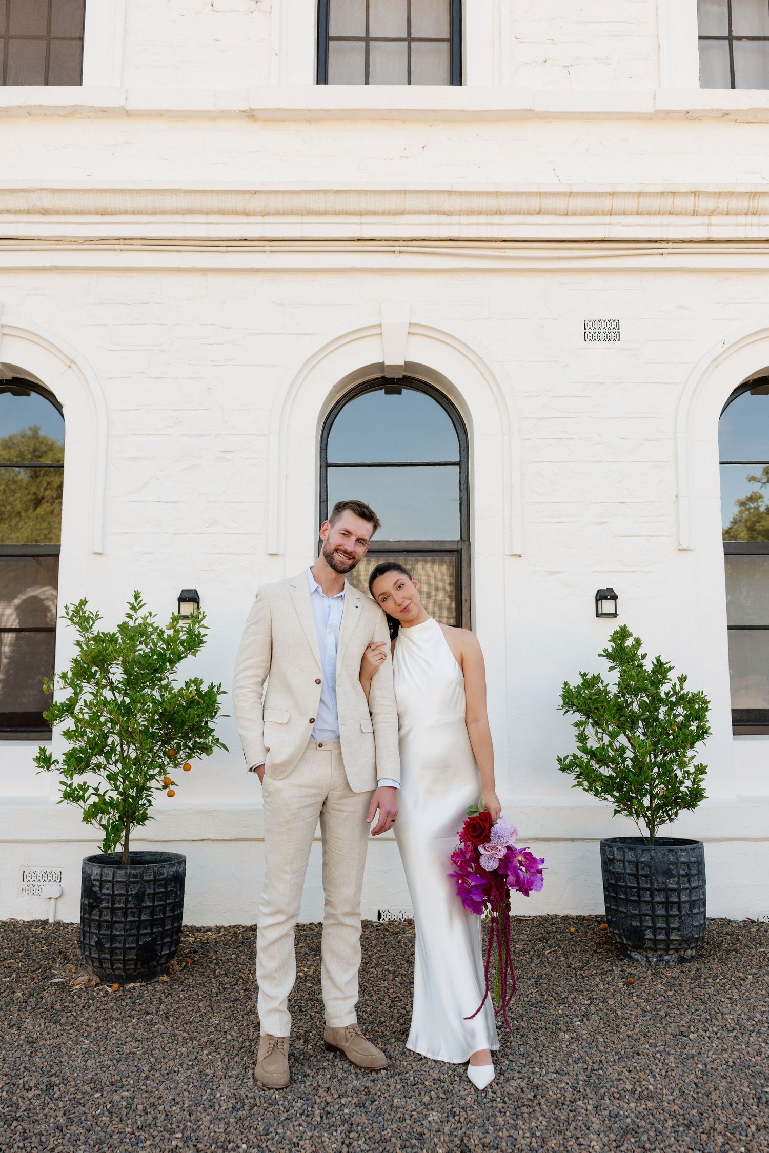 Bride and groom pose before white heritage building, bride in satin gown holding purple bouquet, groom in beige suit.