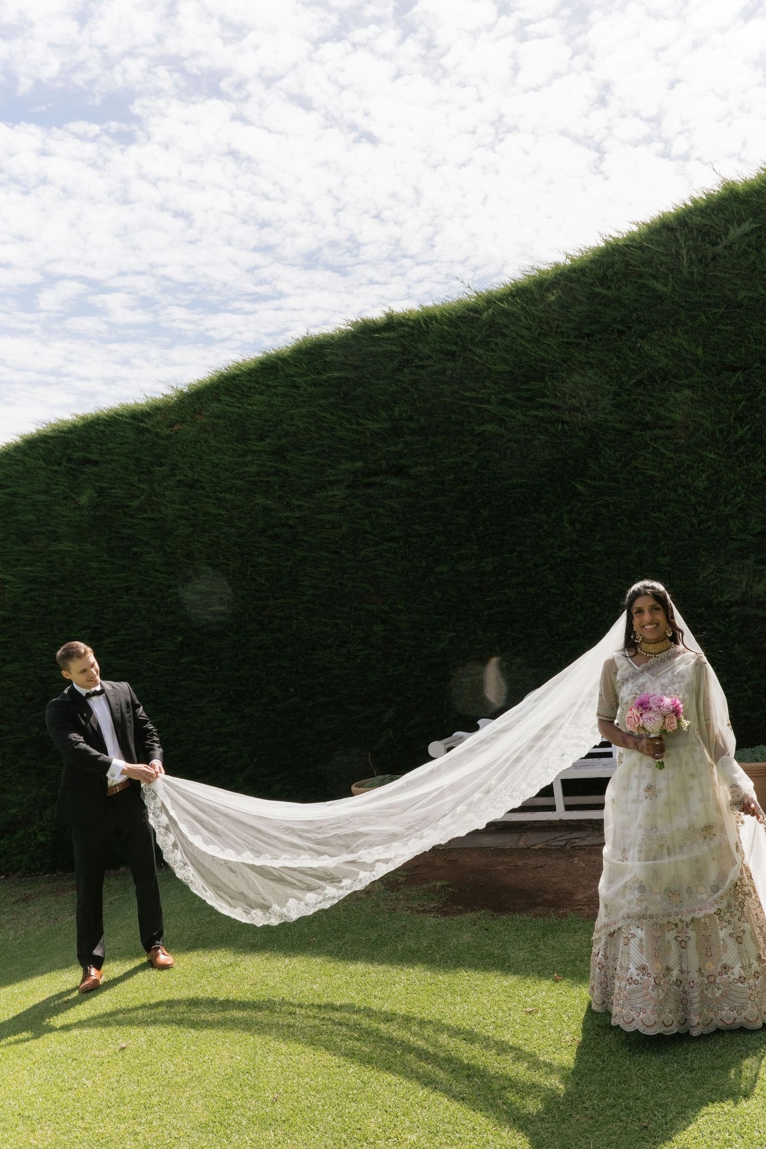 Groom holds flowing white lace veil as bride in Indian lehenga stands on manicured garden lawn.