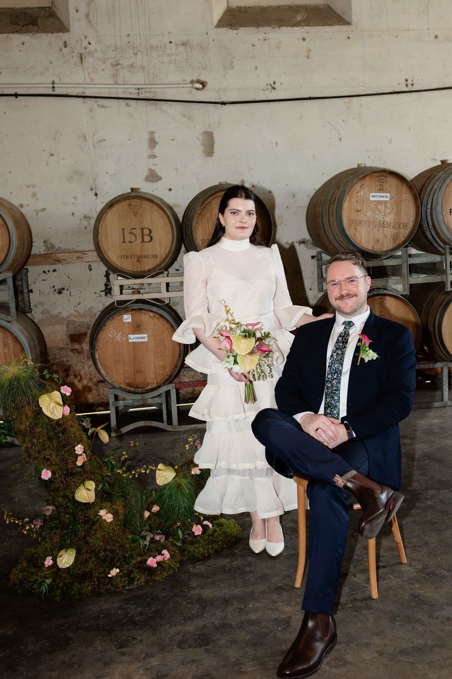 Bride and groom pose among oak wine barrels in rustic winery wedding venue.