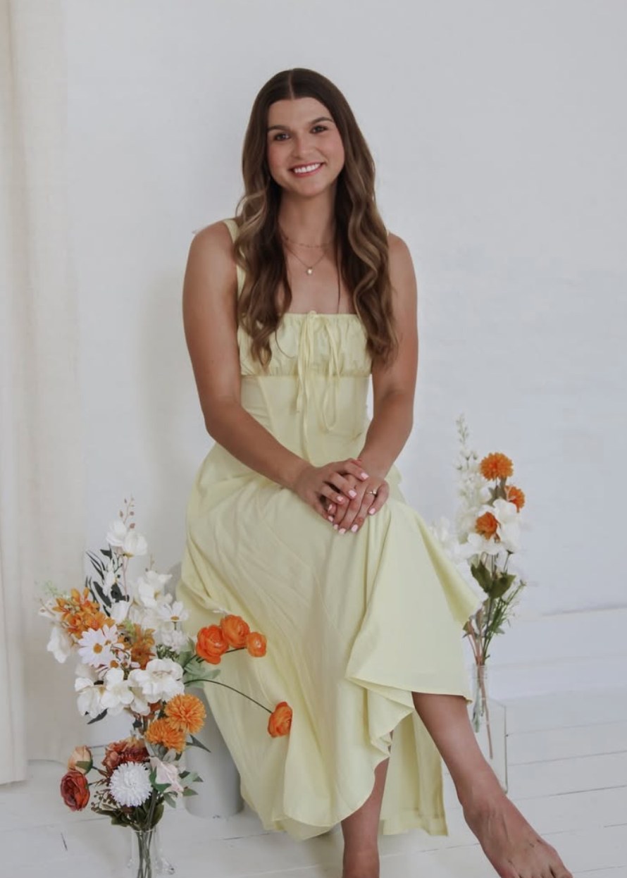 Woman in yellow midi dress sitting among floral arrangements against white background.