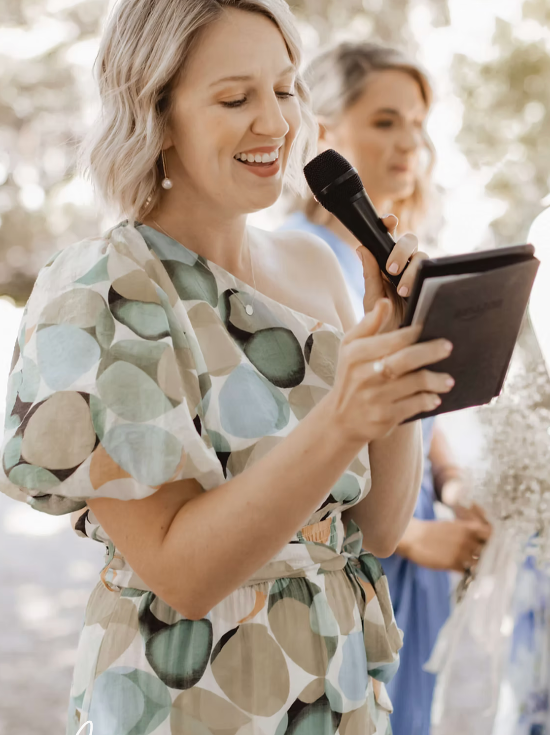Woman in floral dress smiling while holding microphone and tablet outdoors at a wedding ceremony.