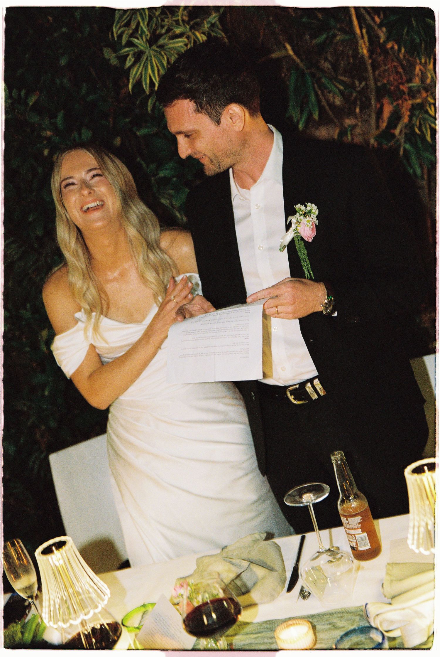 Bride and groom laughing together while reading vows at outdoor wedding reception.