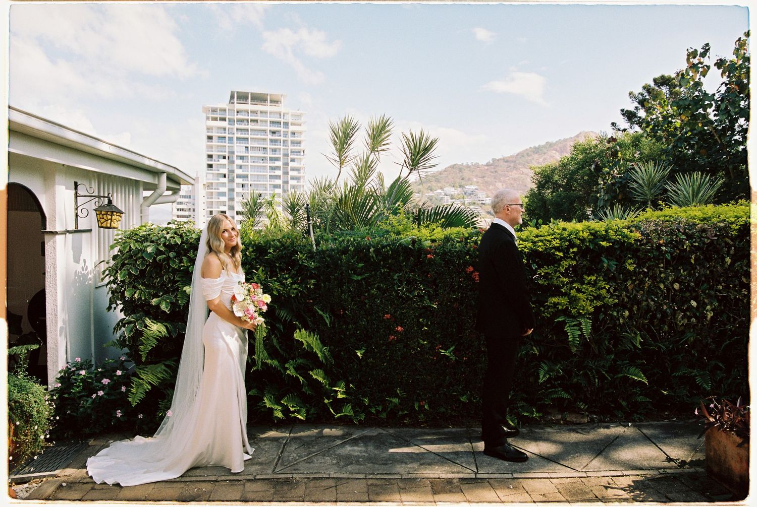 Bride in white gown with veil holding bouquet stands beside groom in garden with mountain view.