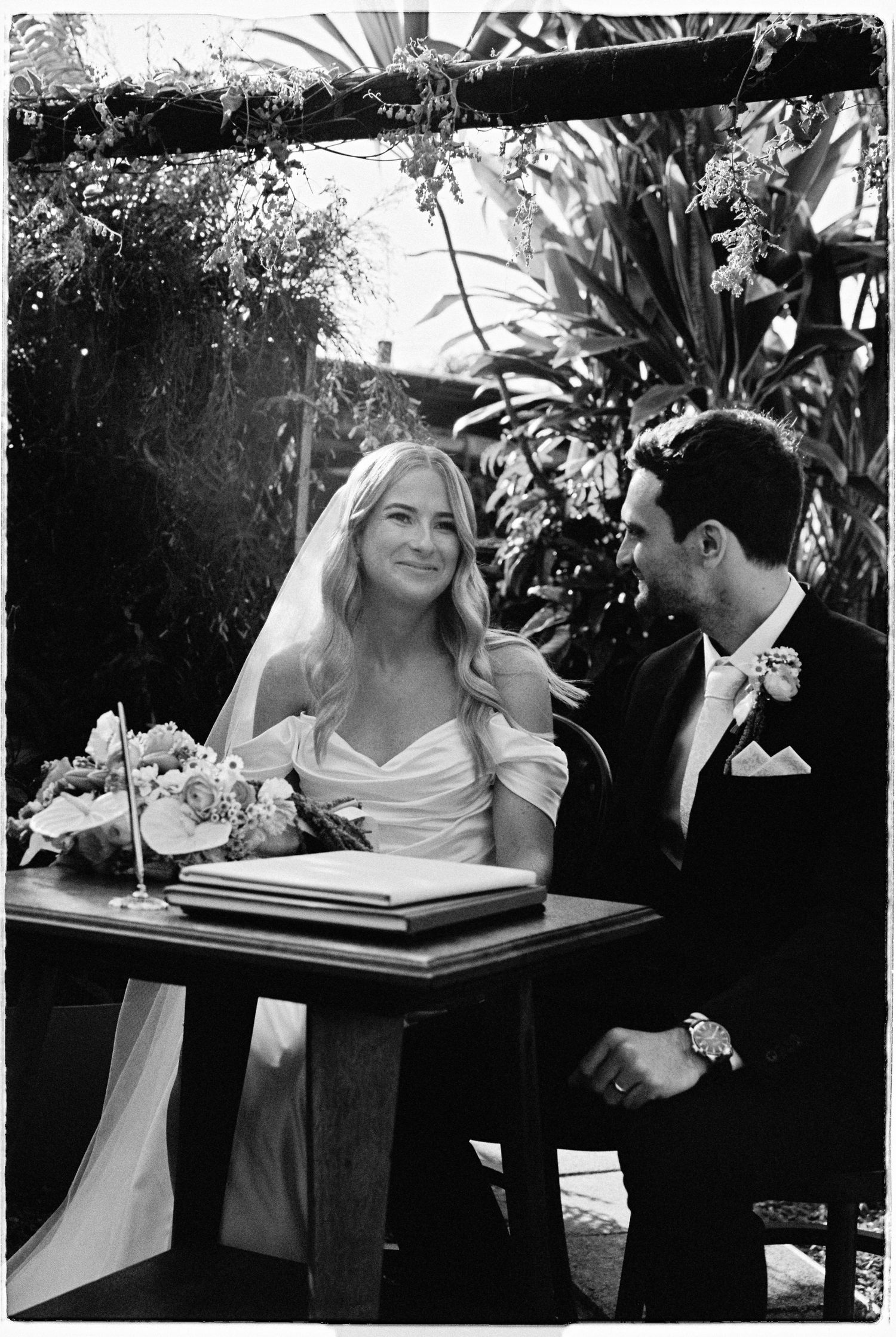Black and white photo of smiling bride and groom signing wedding register at outdoor ceremony table.
