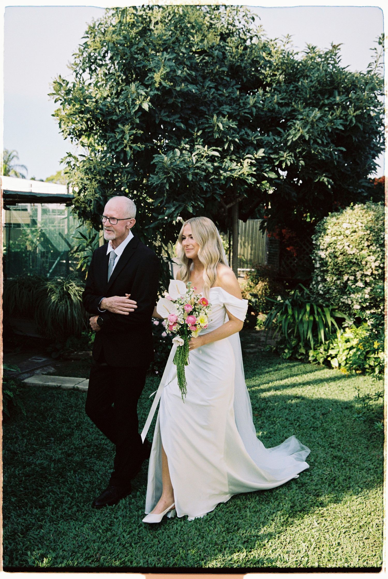 Bride in white gown walking with older man in suit through lush green garden.