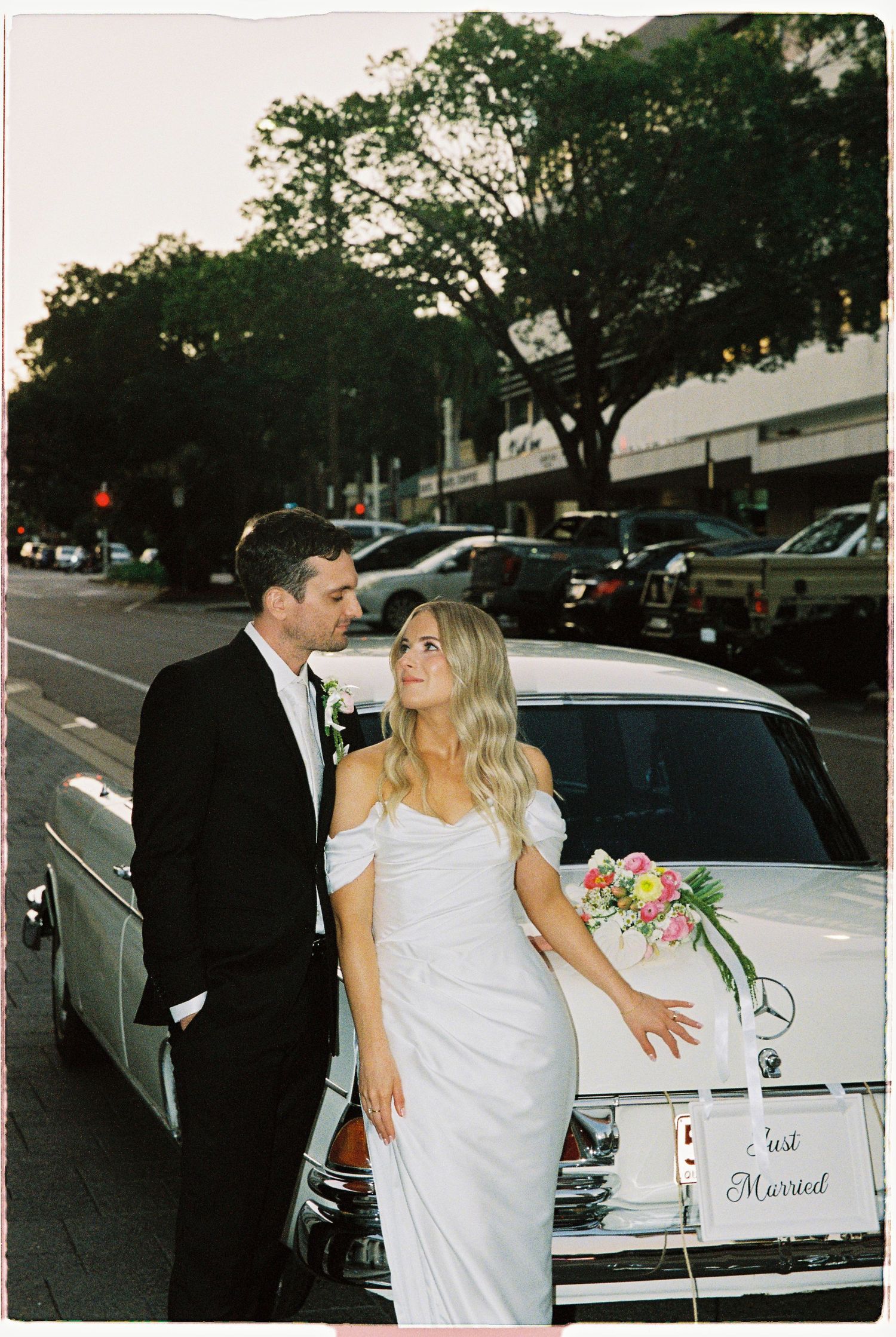 Bride and groom posing beside vintage white Mercedes Just Married car on city street at dusk.