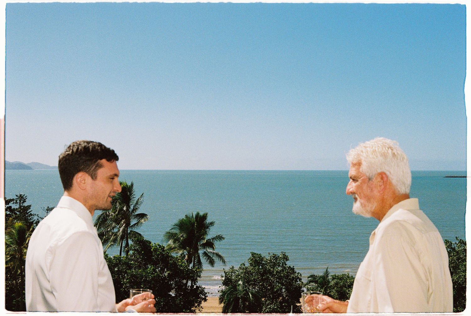 Two men in white shirts converse on a tropical oceanview terrace with palm trees and blue sea backdrop.