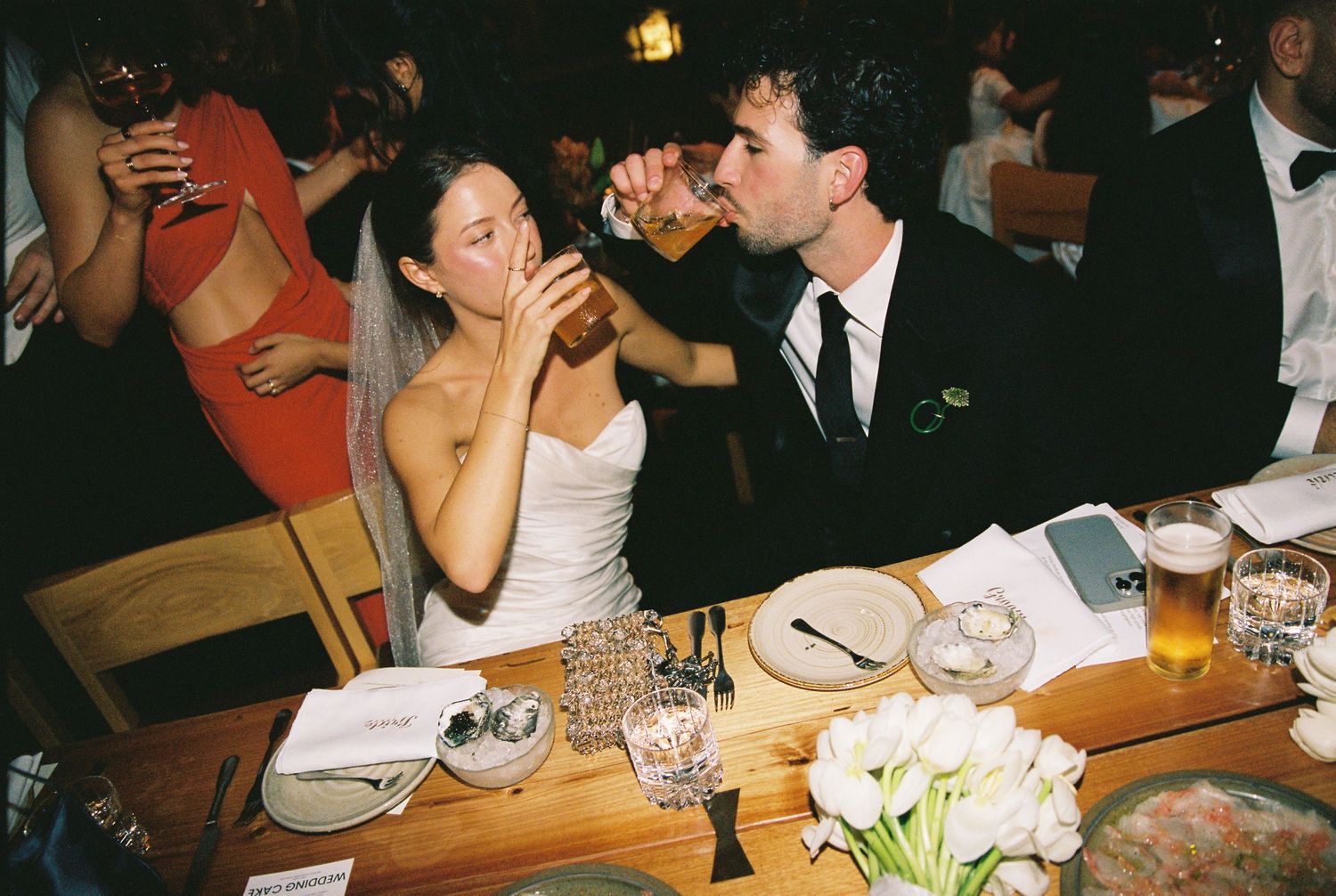 Bride and groom share a playful drink together at their wedding reception dinner table with flowers.