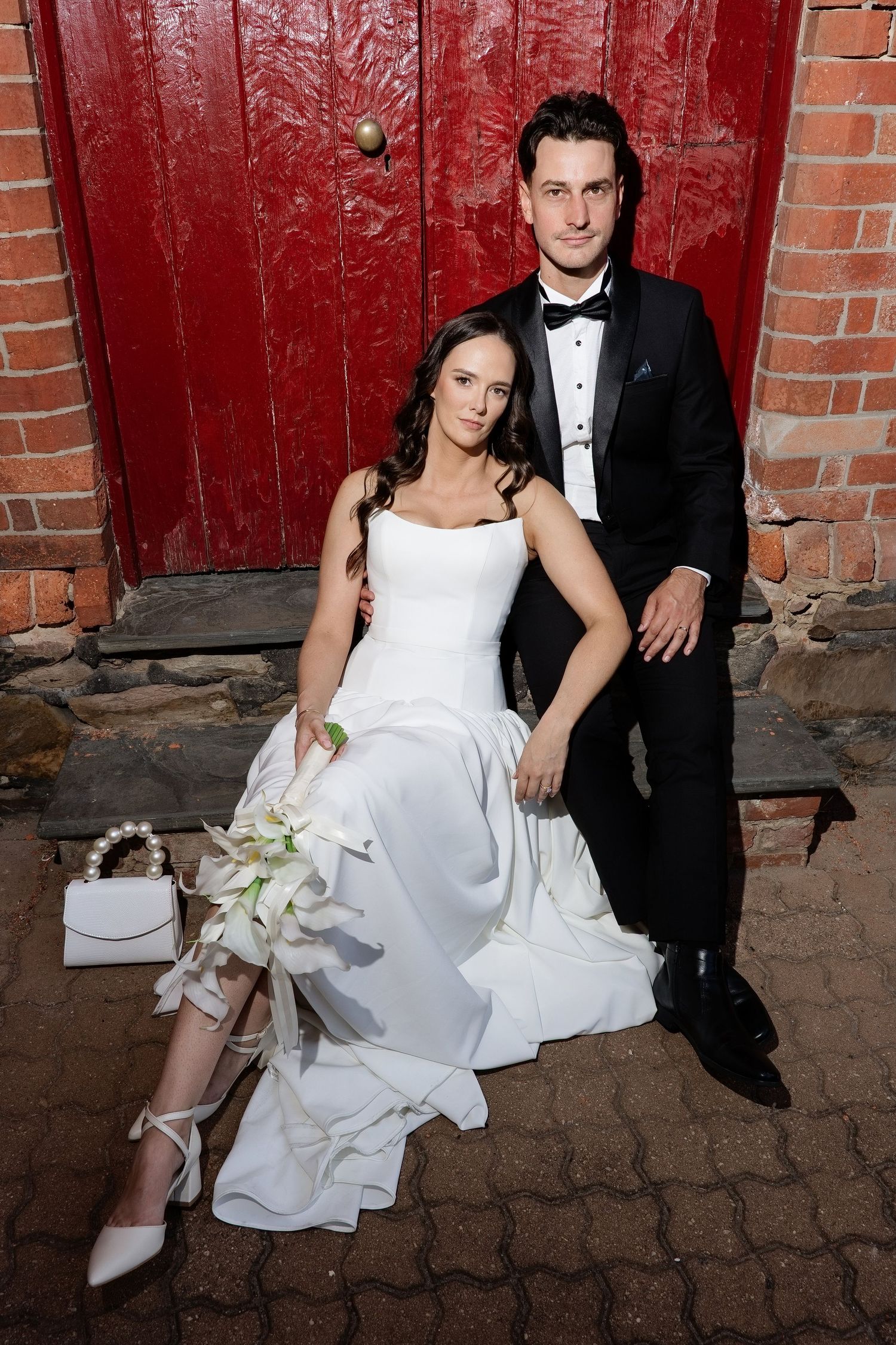 Bride and groom smiling together in modern marble interior, groom seated in green chair, bride holding bouquet.