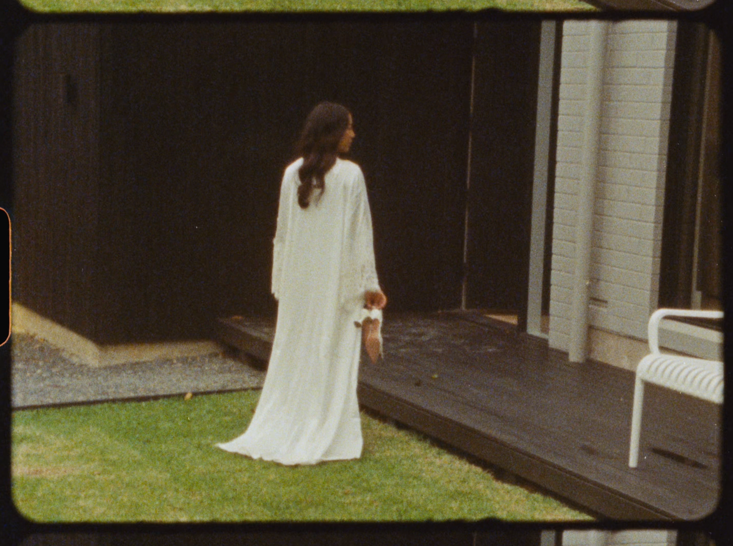 Bride in a flowing white long-sleeve gown standing on a green lawn near a dark wooden building.