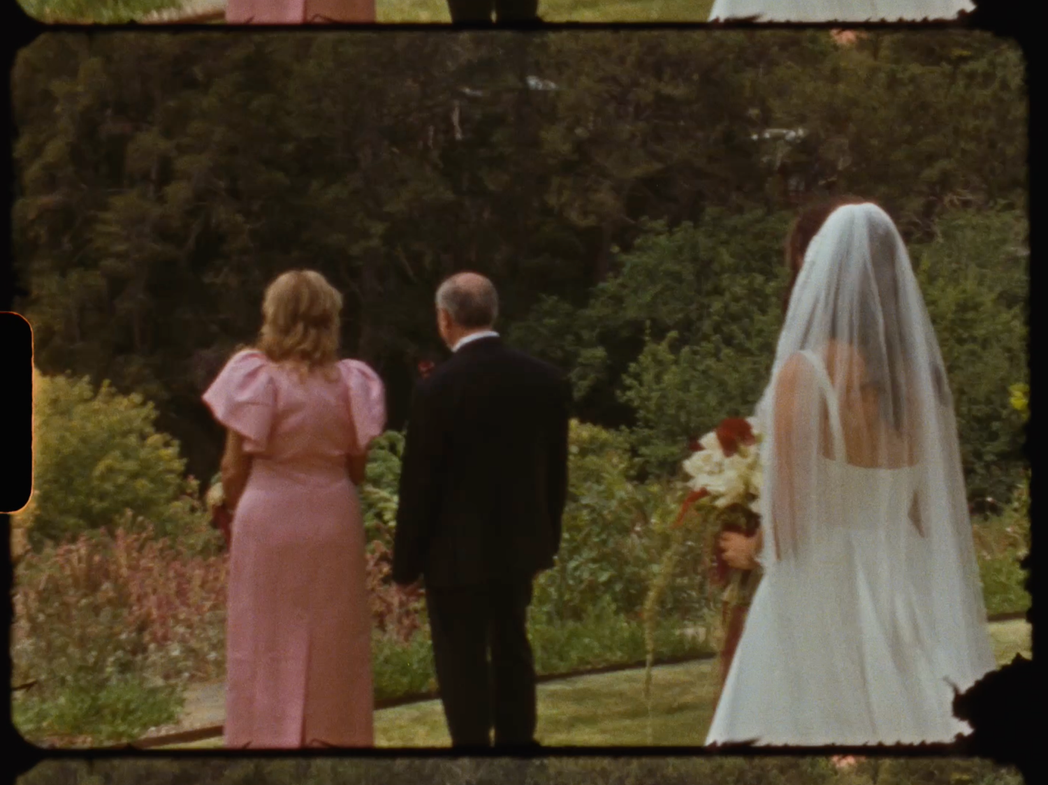 Bride in white gown and veil walks with guests through a lush garden setting.