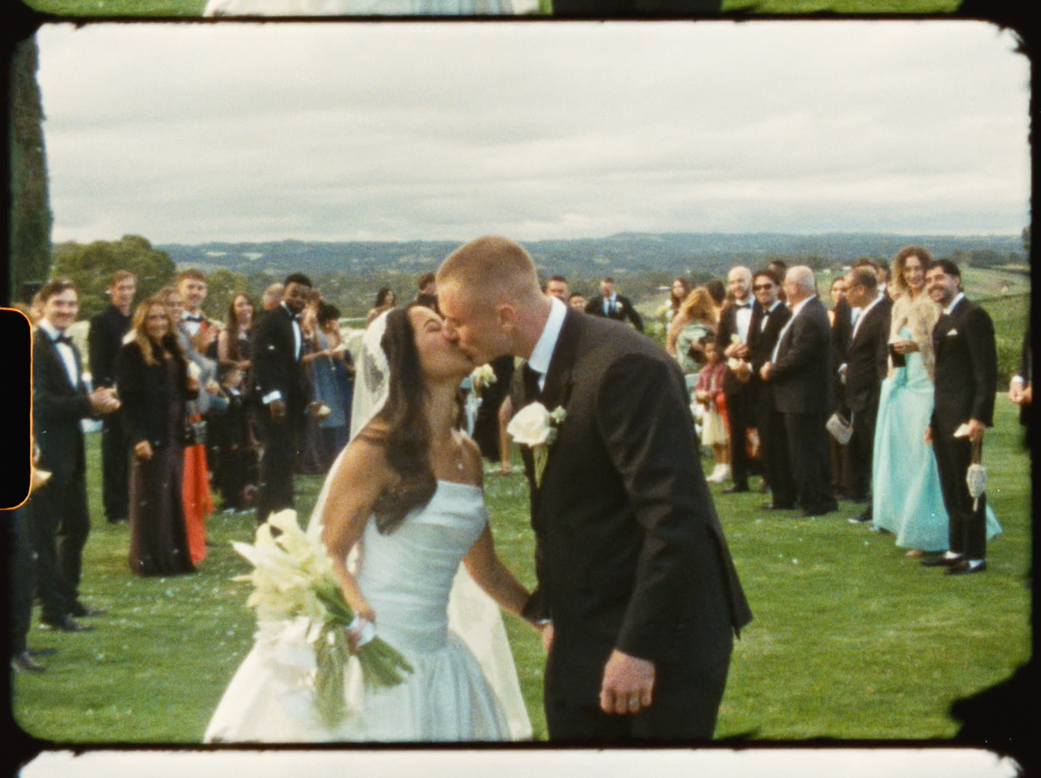 Bride in blue gown and groom kissing at altar surrounded by cheering wedding guests on green lawn.