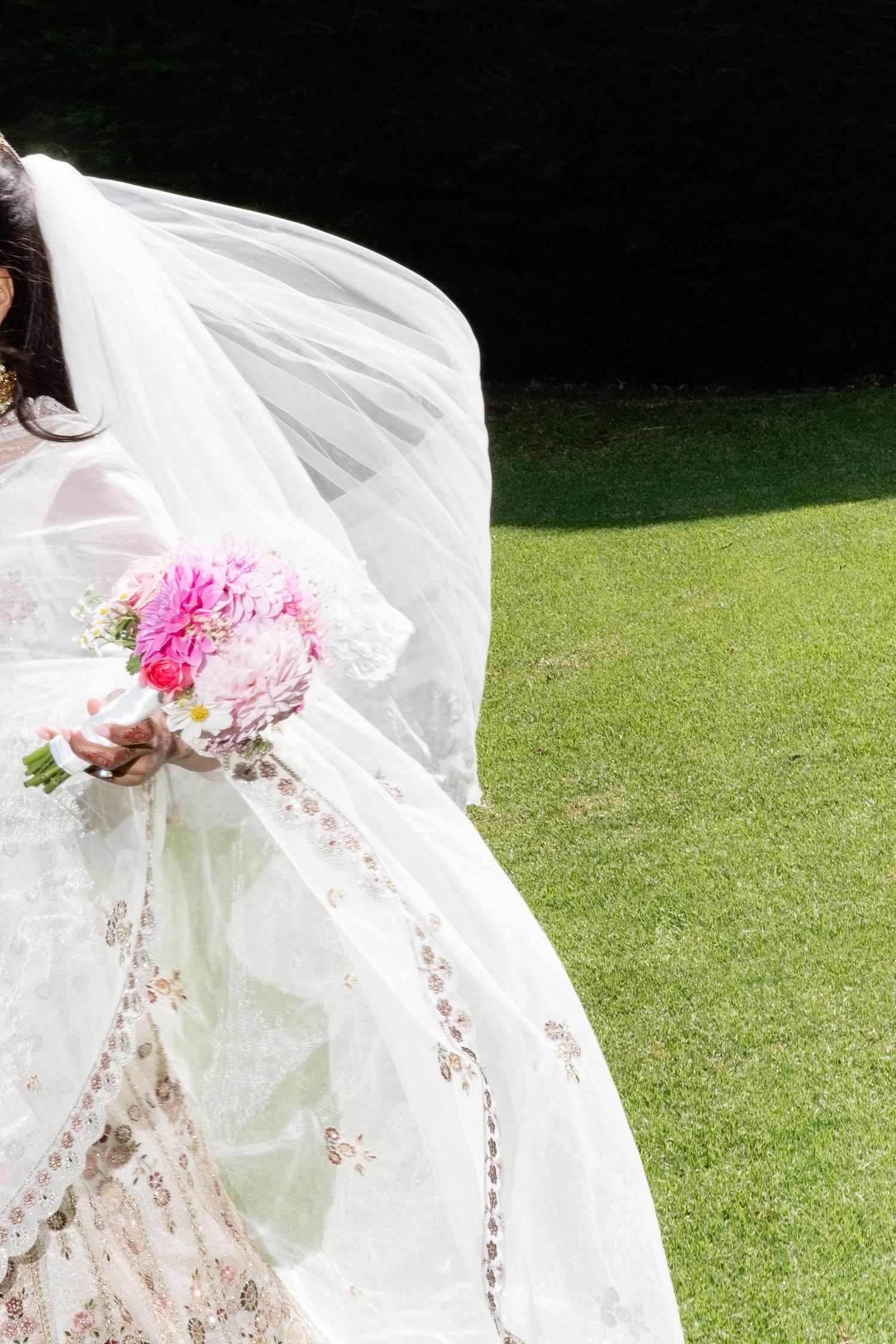 Close-up of bride in floral lehenga with billowing white veil, holding vibrant pink wedding bouquet outdoors.