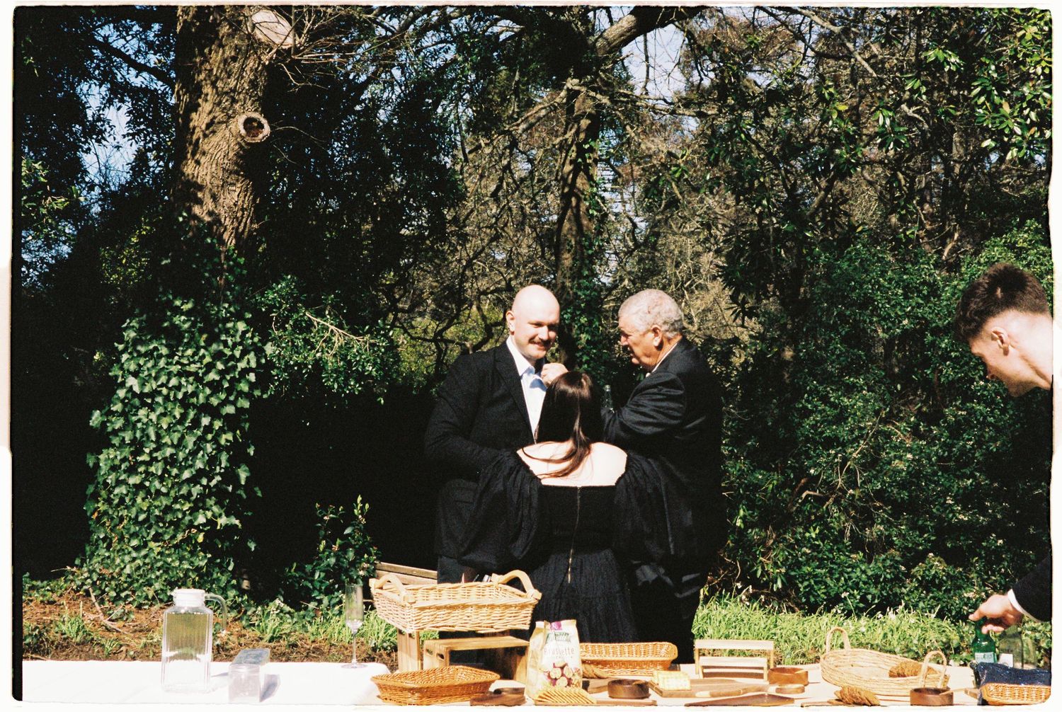 Two men in formal black suits examine items on a table at an outdoor garden wedding celebration.