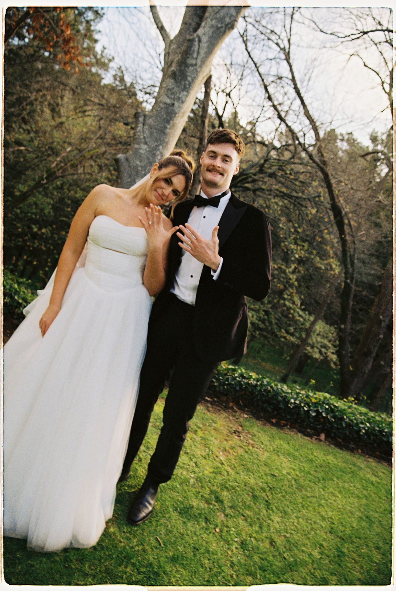 Bride and groom in white gown and black tuxedo pose playfully on green lawn in garden setting.