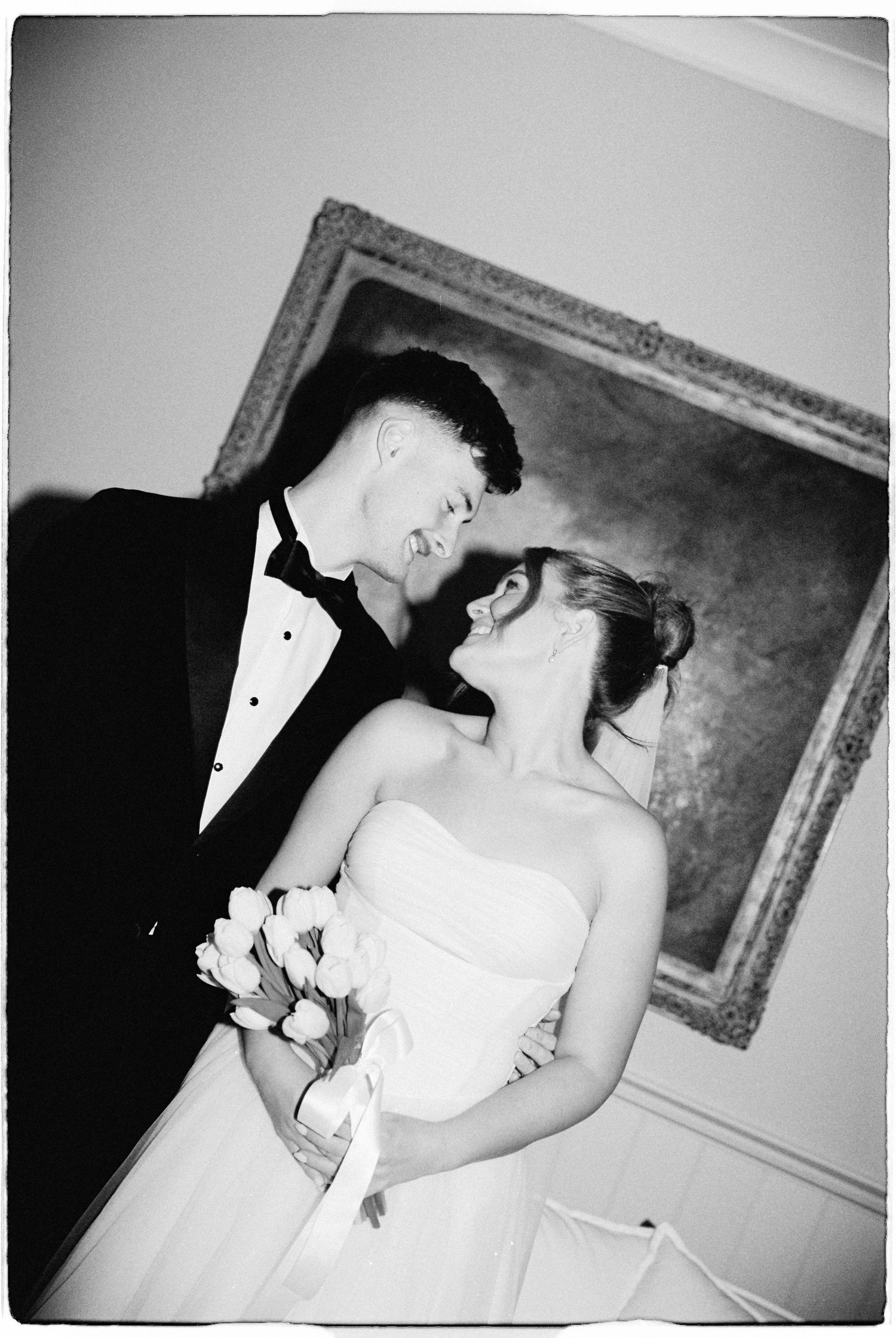 Black and white wedding photo of smiling bride holding tulips and groom in tuxedo sharing romantic moment indoors.