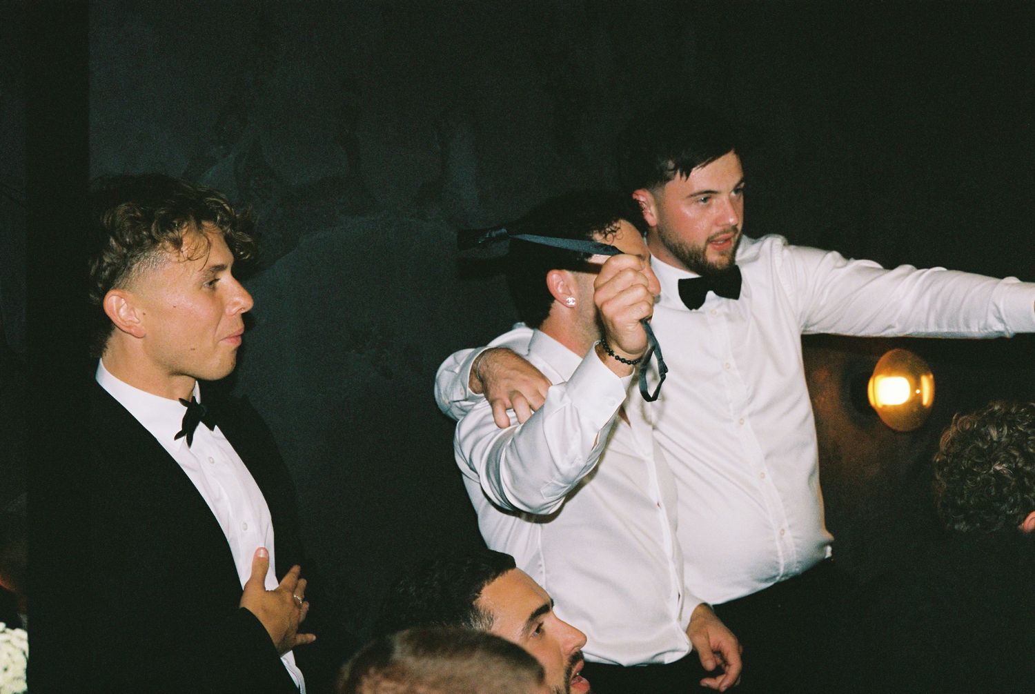 Two men in tuxedos pose playfully with a microphone at a wedding reception.