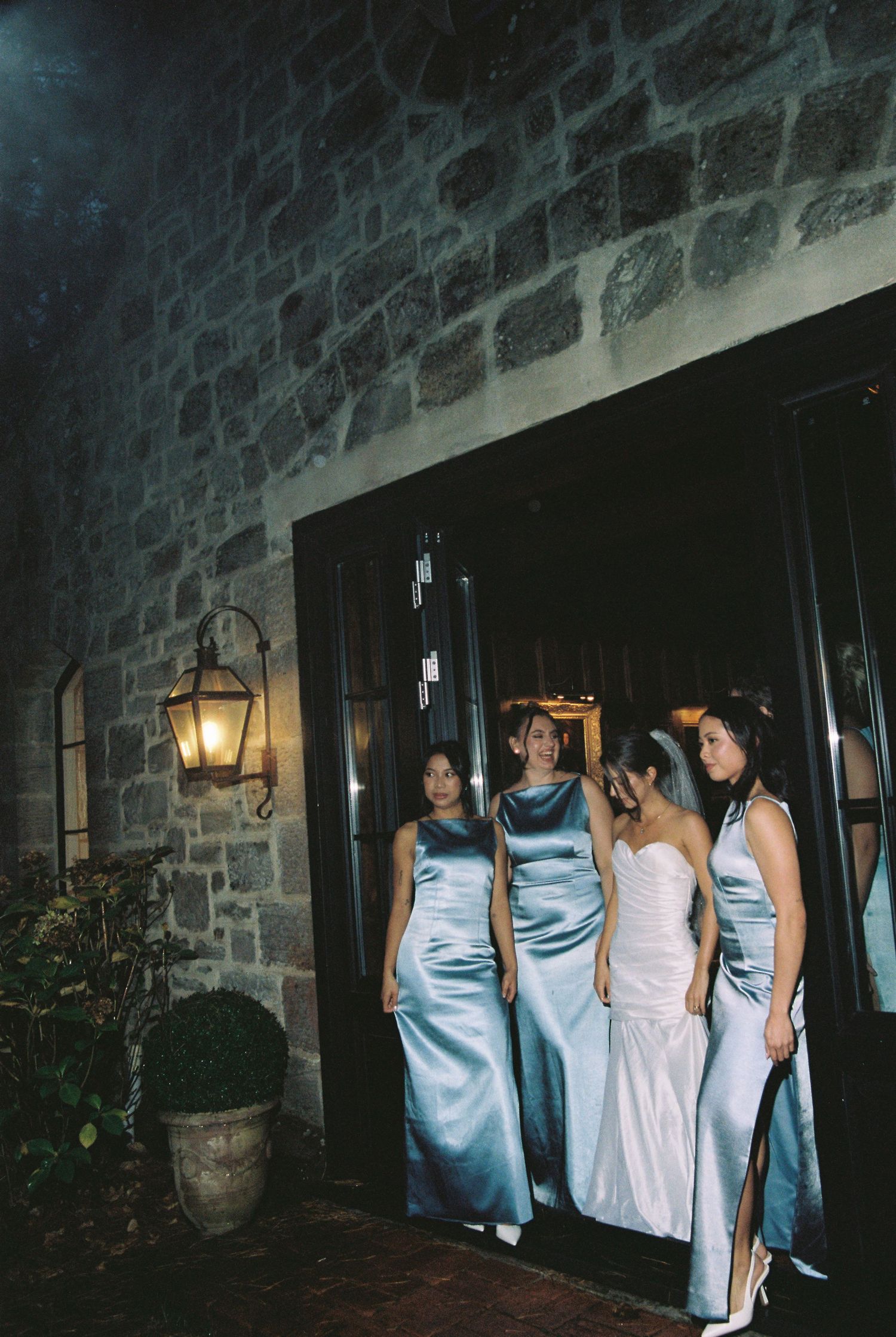 Bride in white gown with bridesmaids in blue satin dresses exiting stone building at night.