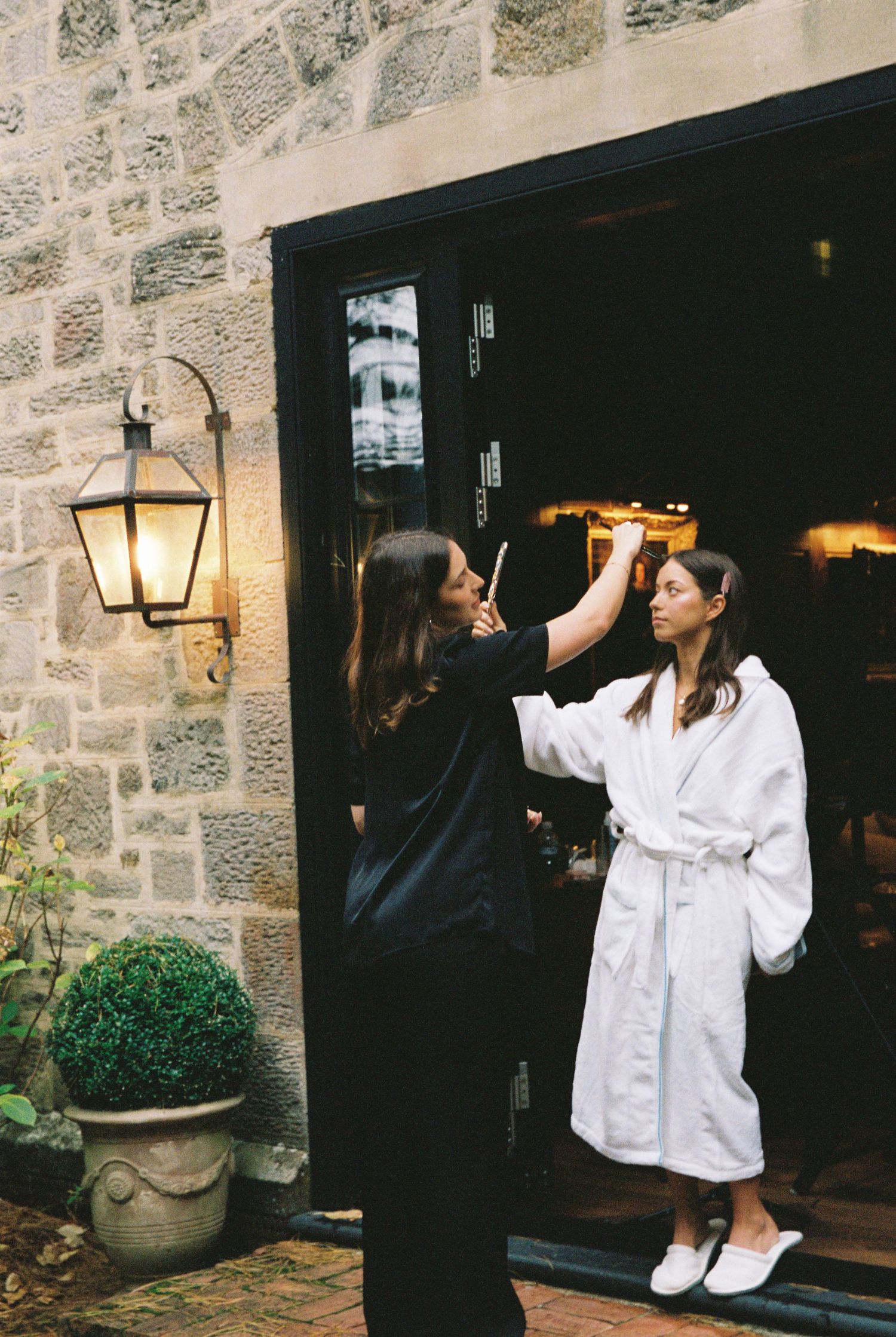 Makeup artist applying makeup to bride in white robe outside stone building entrance.