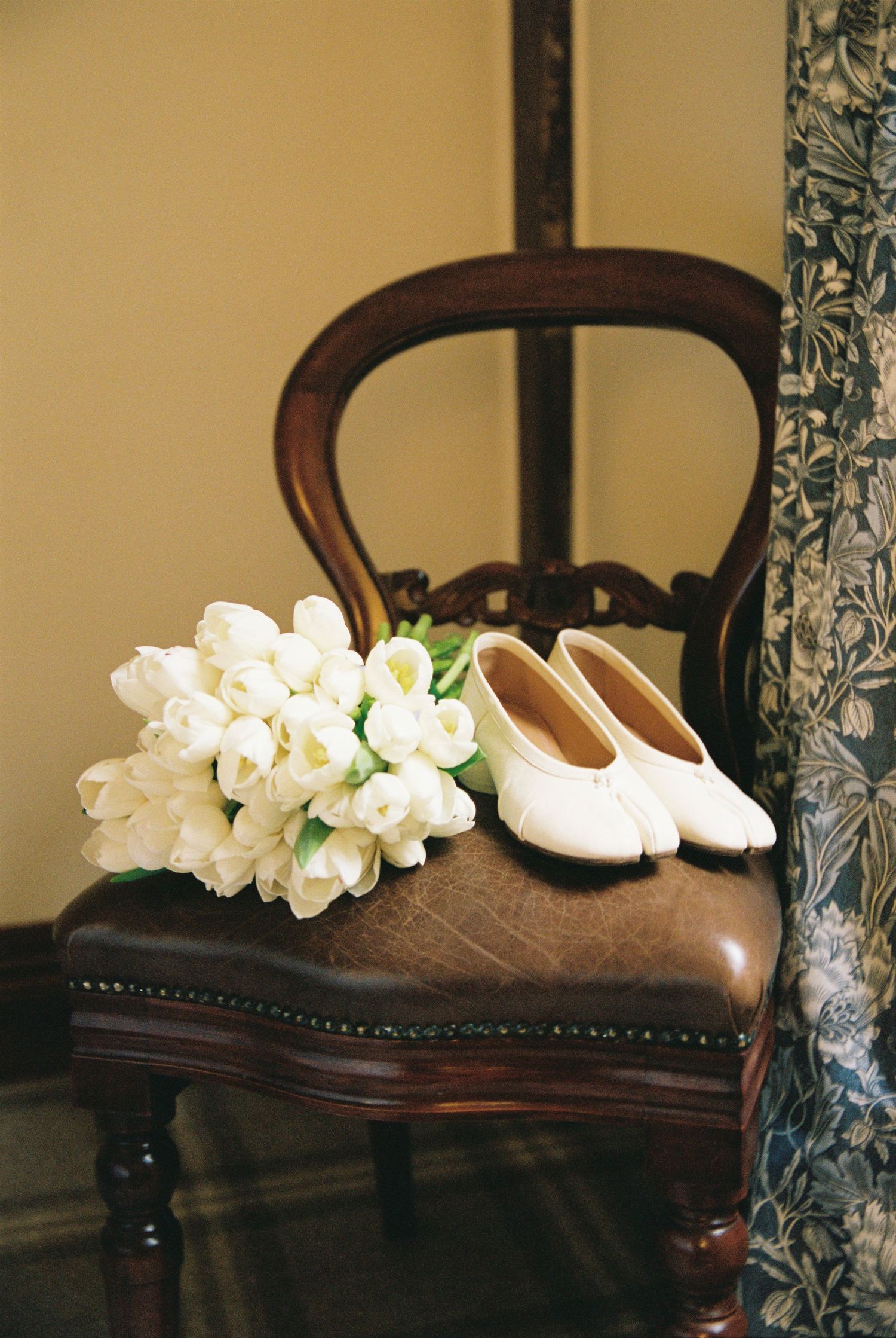 White tulip bridal bouquet and ivory ballet flats rest on vintage wooden chair.