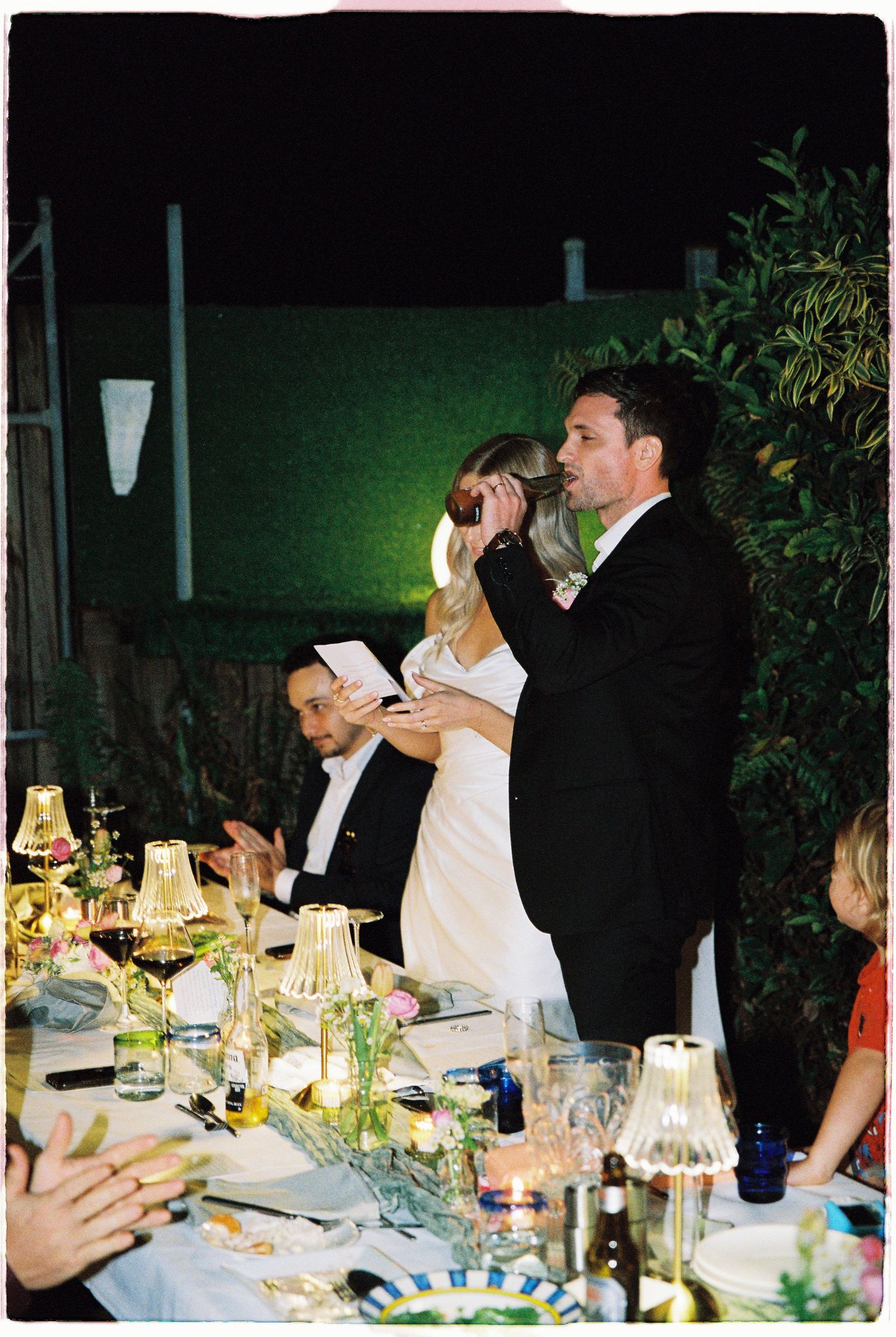 A bride and groom give a speech together at a candlelit outdoor wedding reception dinner.