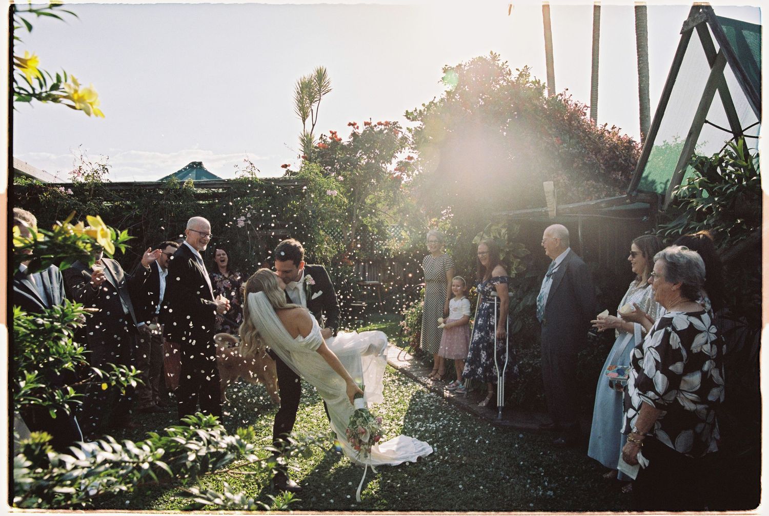 Wedding guests celebrate with confetti as bride and groom embrace in sunlit lush garden ceremony.