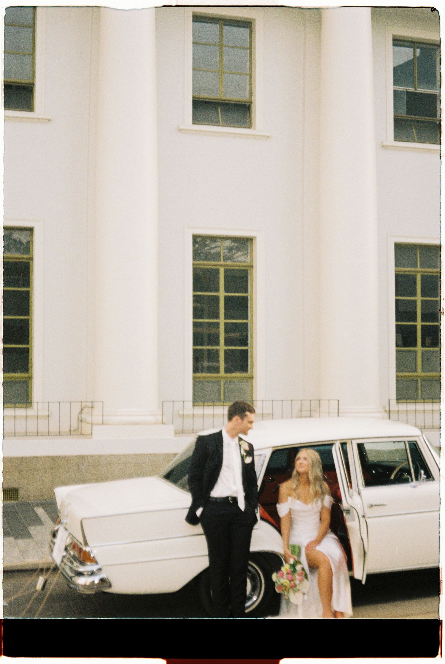 Bride in white gown and groom in black tuxedo posing beside a vintage white car outside a grand building.