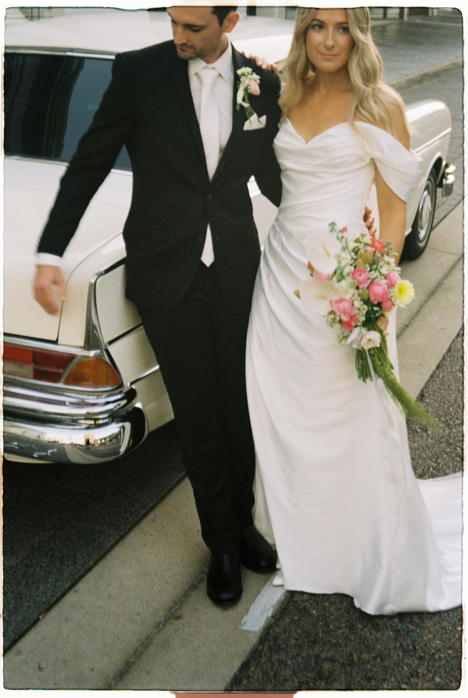 Wedding couple walks beside a classic white vintage car, bride in draped off-shoulder gown carrying a bright pink bouquet.