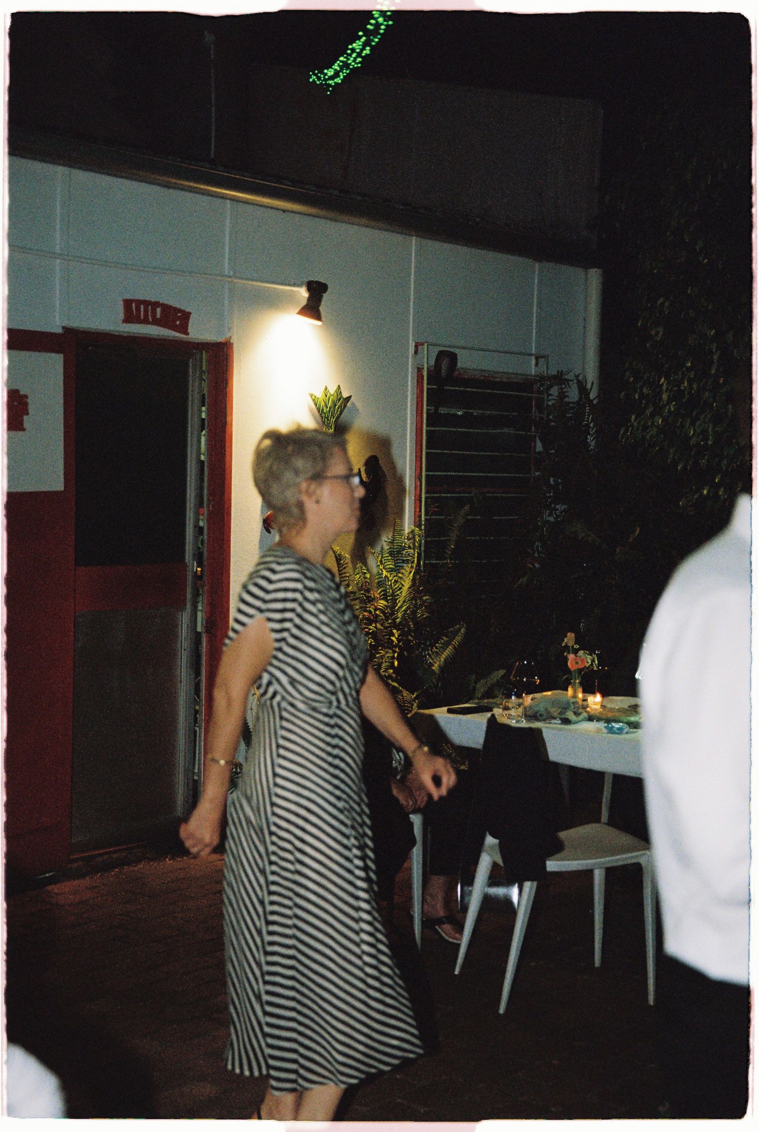 Older woman in striped dress walking at outdoor evening wedding venue near illuminated doorway.