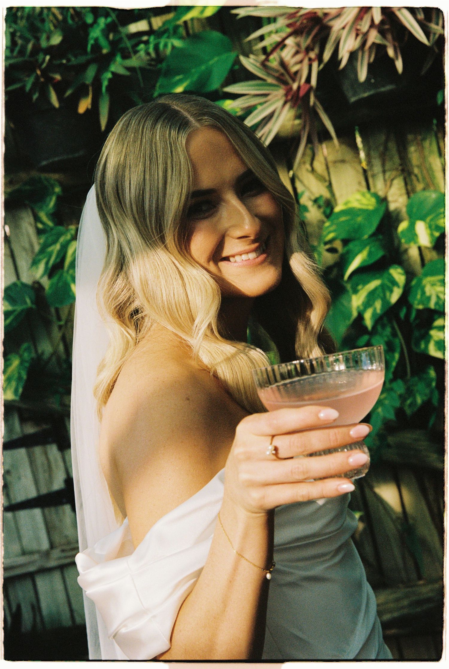 Smiling bride in white off-shoulder gown and veil holds pink cocktail amid lush tropical greenery.