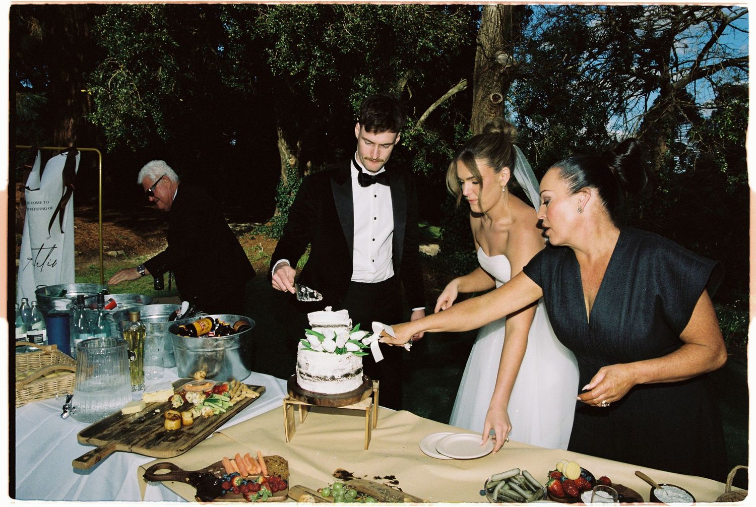 Bride and groom cutting their wedding cake outdoors at night with a guest assisting.