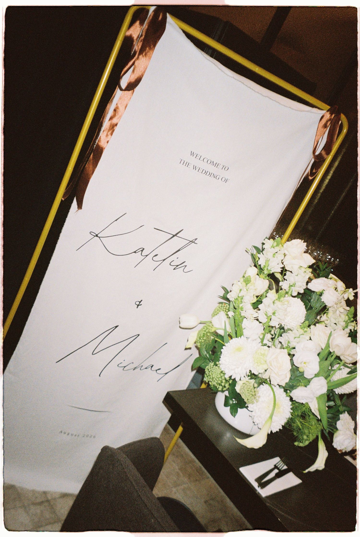 Welcome sign for the wedding of Kaitlin and Michael displayed beside white floral arrangement.